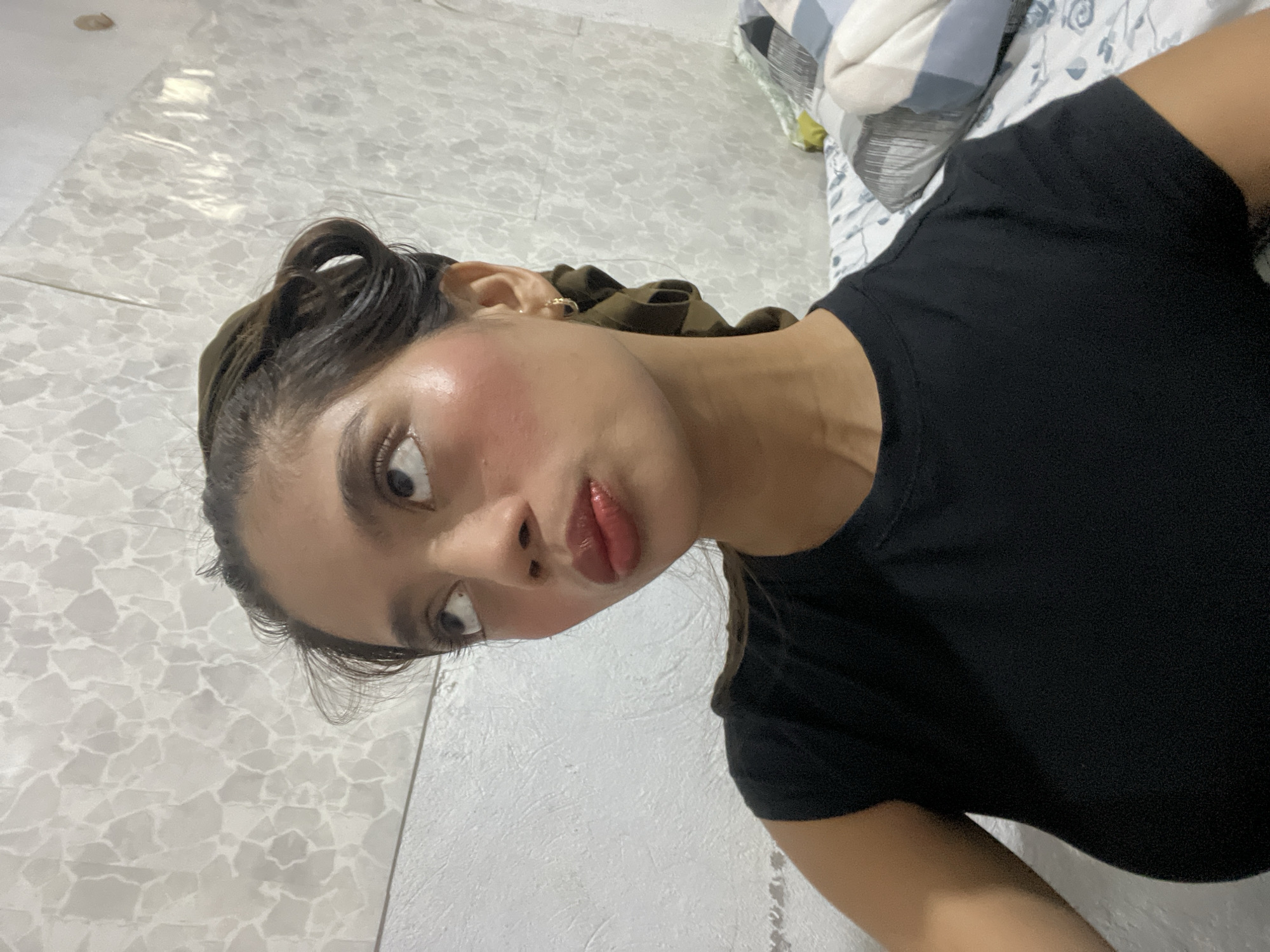 A high-angle, portrait-style selfie captures a young woman lying down in what appears to be an indoor setting, possibly a bedroom in Davao City, Philippines. She has dark hair, partially tied back with a dark green or brown scrunchie, and wears a small gold hoop earring. Her black t-shirt covers her torso.
The woman's face is angled towards the viewer, featuring wide, dark eyes and slightly pursed, glossy lips with a reddish tint. A noticeable rosy blush is visible on her cheeks, possibly from makeup or a natural flush. Her expression conveys a sense of playful curiosity or surprise.
The background includes a patterned bedsheet, white with grey floral or swirling designs, and two pillows—one with blue and white stripes, and another with a similar floral pattern to the sheet. A light-toned floor or wall, characterized by a mosaic-like pattern of irregular grey and white tiles, occupies the bottom half and left side of the frame, contributing to the casual home environment. There is no visible text in the image.
