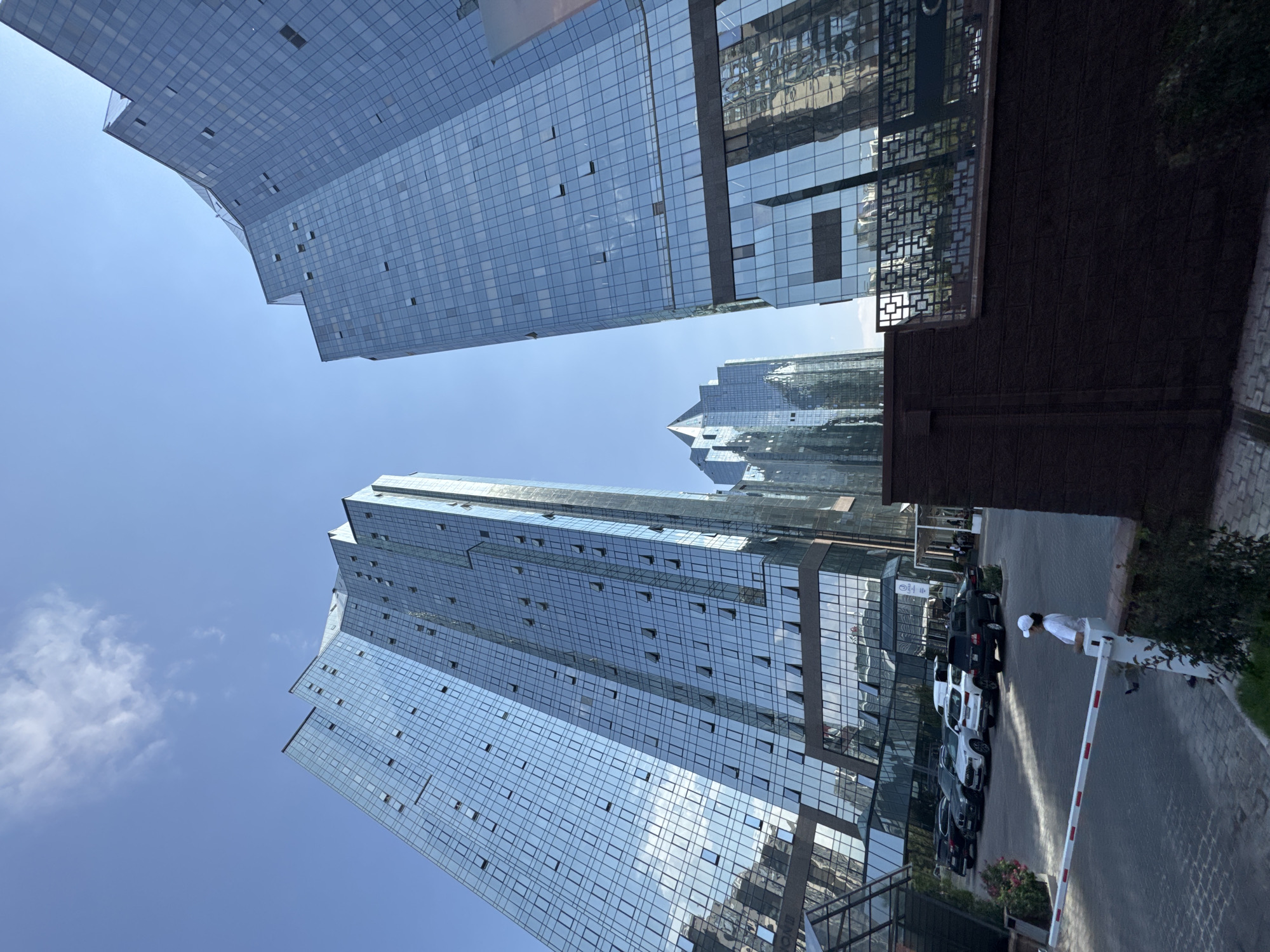 In Almaty, Kazakhstan, a daytime, wide-angle, low-angle shot captures a modern architectural ensemble of glass-facade skyscrapers. The sky is clear blue with a few wispy clouds. In the foreground, a white retractable barrier arm is positioned across a paved driveway. A person wearing a white cap and shirt stands to the right of the barrier arm, facing away from the camera. To the left of the barrier, several cars are parked in a lot. Behind the barrier and parking area, the lower sections of the skyscrapers are visible, featuring dark brown brick walls and decorative metal railings. The dominant subjects are the towering glass buildings, which reflect the sky and surrounding structures.