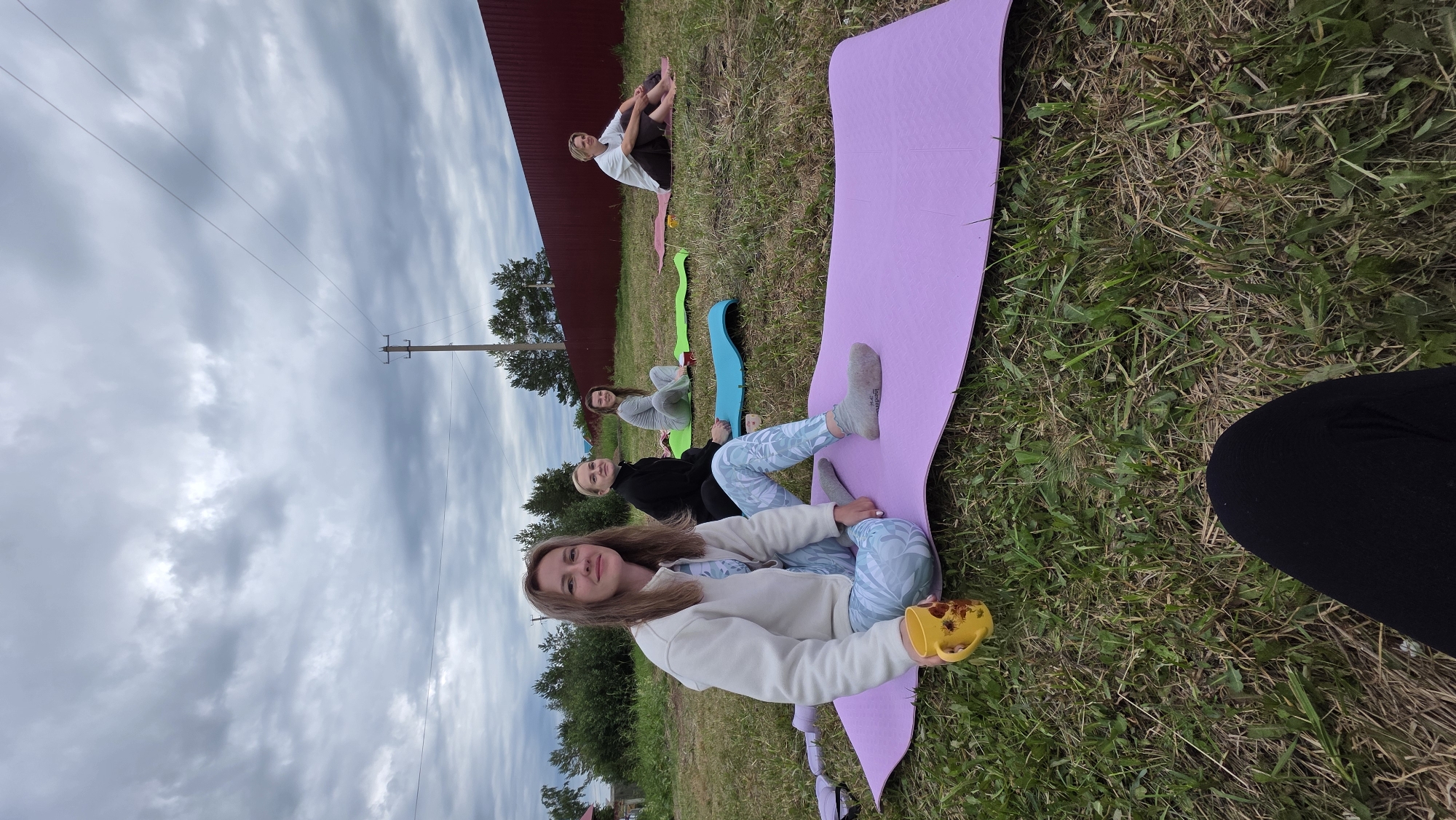 In the city of Kupoyarovo, Russia, a group of four women are seen sitting on yoga mats outdoors on a cloudy day. The woman in the foreground, wearing a white zip-up hoodie and light blue patterned leggings, is holding a yellow mug with a sunflower design. She has light brown hair and is looking towards the camera. Behind her, three other women are seated on yoga mats of various colors (green, blue, and pink). The woman in the black top and dark pants is in the middle ground. Further back, a woman in a light grey top and light pants is seated on a green mat, and another woman in a white top and dark pants is on a pink mat. The women appear to be engaged in a yoga session or a similar outdoor activity. The environment is grassy, with trees and a red fence visible in the background. A telephone pole with wires is also present in the scene. The overall atmosphere suggests a casual, outdoor gathering.