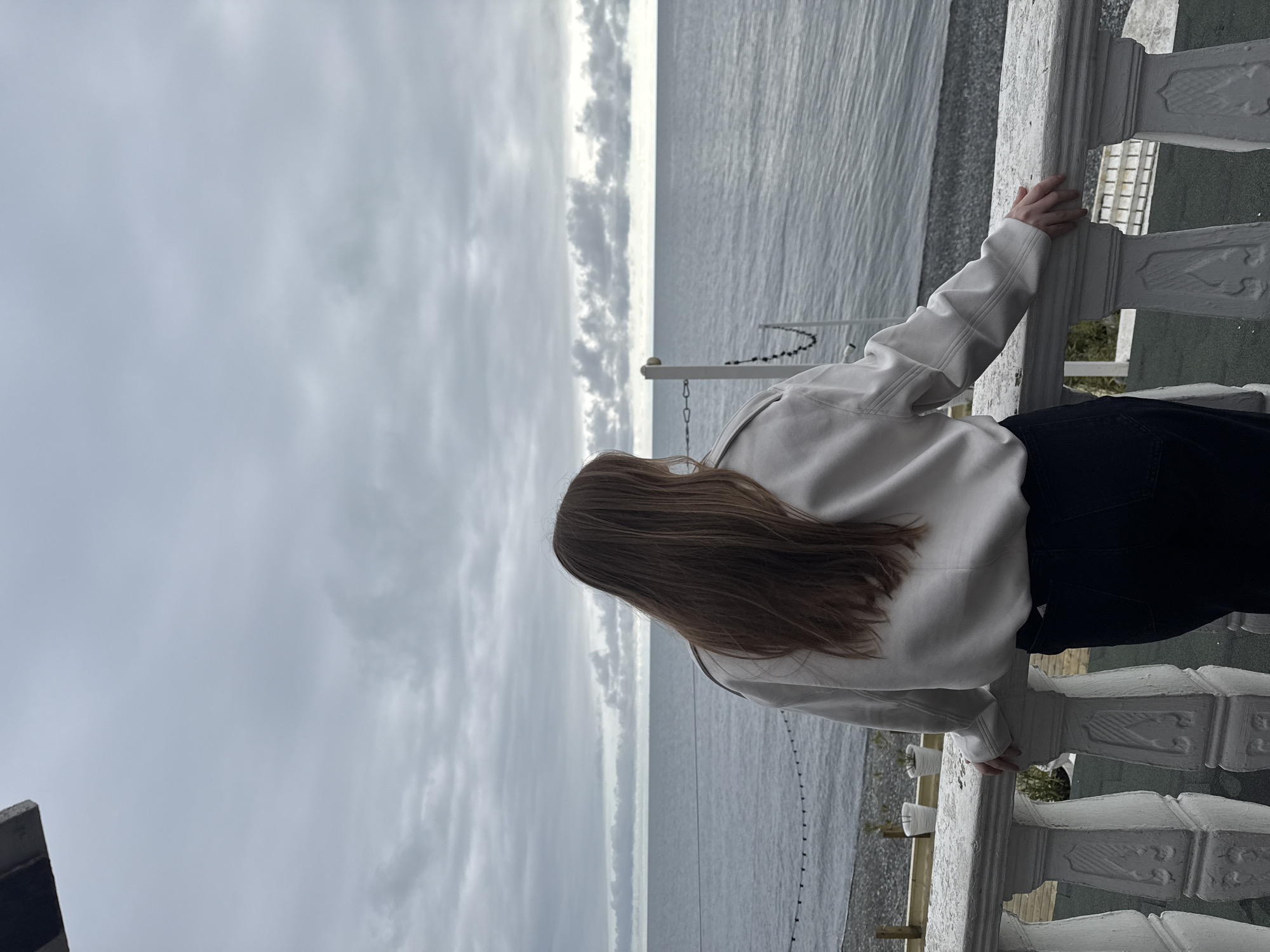 A lone individual is depicted from behind, centered on a weathered white stone balustrade, overlooking the Black Sea in Sochi, Russia. The subject has long, straight, brown hair extending to their mid-back and is dressed in a light-colored, off-white long-sleeved top or jacket and dark trousers. Their hands rest upon the top rail of the balustrade, which features classical-style carved balusters showing signs of wear. Below the balustrade, a dark pebble beach is visible, where two white potted plants are positioned on the left, and a white vertical post with a black chain draped from it is observed on the right. The calm, grey-blue water of the Black Sea extends to a flat horizon under a uniformly grey, cloudy sky.