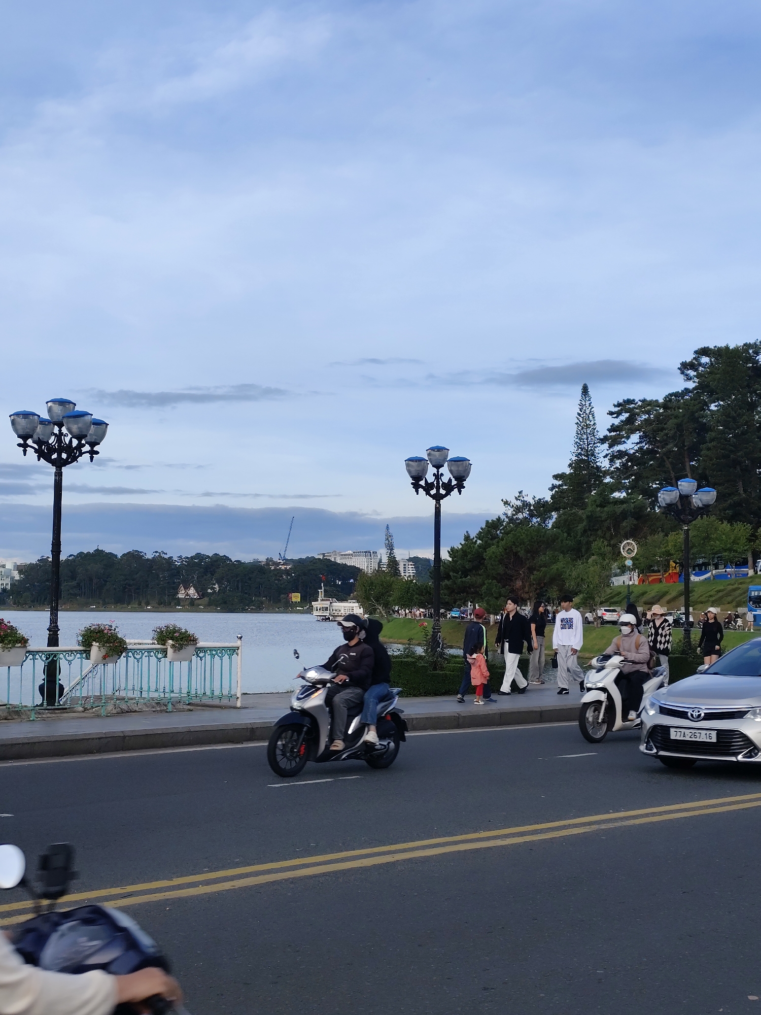 The image captures a street scene in Da Lat, Vietnam, near Xuan Huong Lake. A paved road with double yellow lines runs across the foreground. On the road, a silver Toyota sedan with license plate "77A-267.16" is traveling to the right, and a scooter with two occupants, a driver wearing a mask and a passenger, is also moving to the right. A third scooter, only partially visible on the left, is facing away from the camera.
To the left of the road, a walkway is bordered by a decorative fence with planters. Beyond the walkway is the expanse of Xuan Huong Lake, with trees and buildings lining its far shore. A distinctive lamppost with four lamps stands prominently on the walkway, and another similar lamppost is visible further down.
On the walkway, several pedestrians are strolling. Among them are a group of young people, some wearing casual attire, and a child in a pink jacket. In the background, the landscape is dominated by lush green trees, a prominent tall pine tree, and some urban structures, including buildings and what appears to be a crane. The sky is overcast with soft clouds.