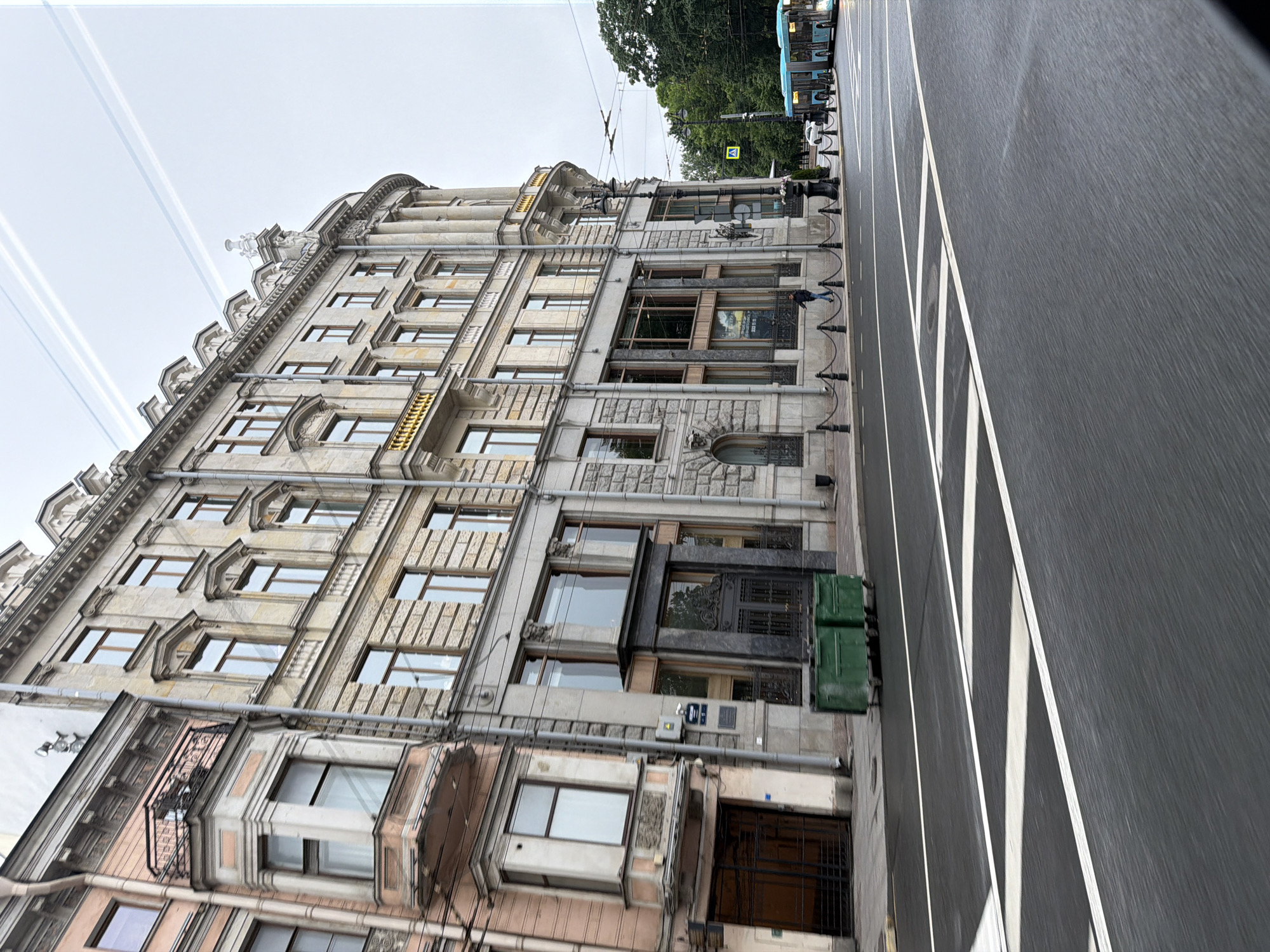 The image shows a view of a street in Saint Petersburg, Russia. The architecture suggests an older, European city. On the left is a light pink building with ornate detailing, including a rounded bay window with a small balcony. To its right is a larger, cream-colored building with multiple floors, characterized by classical architectural elements like columns, decorative moldings, and numerous windows with wooden frames. The street in the foreground is wet and has white lane markings. There are green trash bins on the sidewalk. Further down the street, a light blue bus is visible, along with trees and a pedestrian crossing sign. The sky is overcast with faint contrails from an aircraft. A lone figure is walking on the sidewalk in front of the cream-colored building.