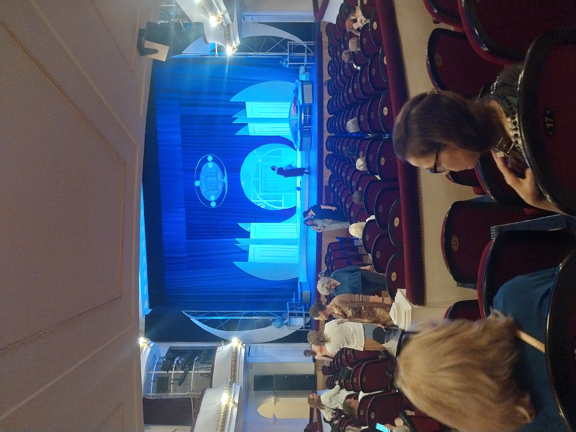 The image shows an audience seated in a theater, facing a brightly lit stage. The theater has rows of red velvet seats, some with numbers visible, such as "17" and "23." The stage is decorated with large, stylized blue and white architectural elements, resembling a futuristic city or abstract art. A blue curtain forms the backdrop. On the stage, a few figures are visible, one dressed in dark attire and another in lighter clothing, appearing to be performers or participants in an event. The lighting on stage is a vivid blue, illuminating the set pieces and creating a dramatic atmosphere.
The audience members are mostly looking towards the stage, suggesting they are attending a performance or presentation. The individuals visible in the foreground and mid-ground are varied in age, with some women and men. One woman in the foreground is looking down at her phone, partially obscured by the seat in front of her. The overall mood seems to be one of attentive observation, though the presence of the phone user suggests a less formal aspect to the event. The scene appears to be indoors, given the artificial lighting and the enclosed nature of the theater. There are no clear indications of time of day or weather conditions. The location context provided is Orenburg, Russia. No other visible text is discernible in the image.