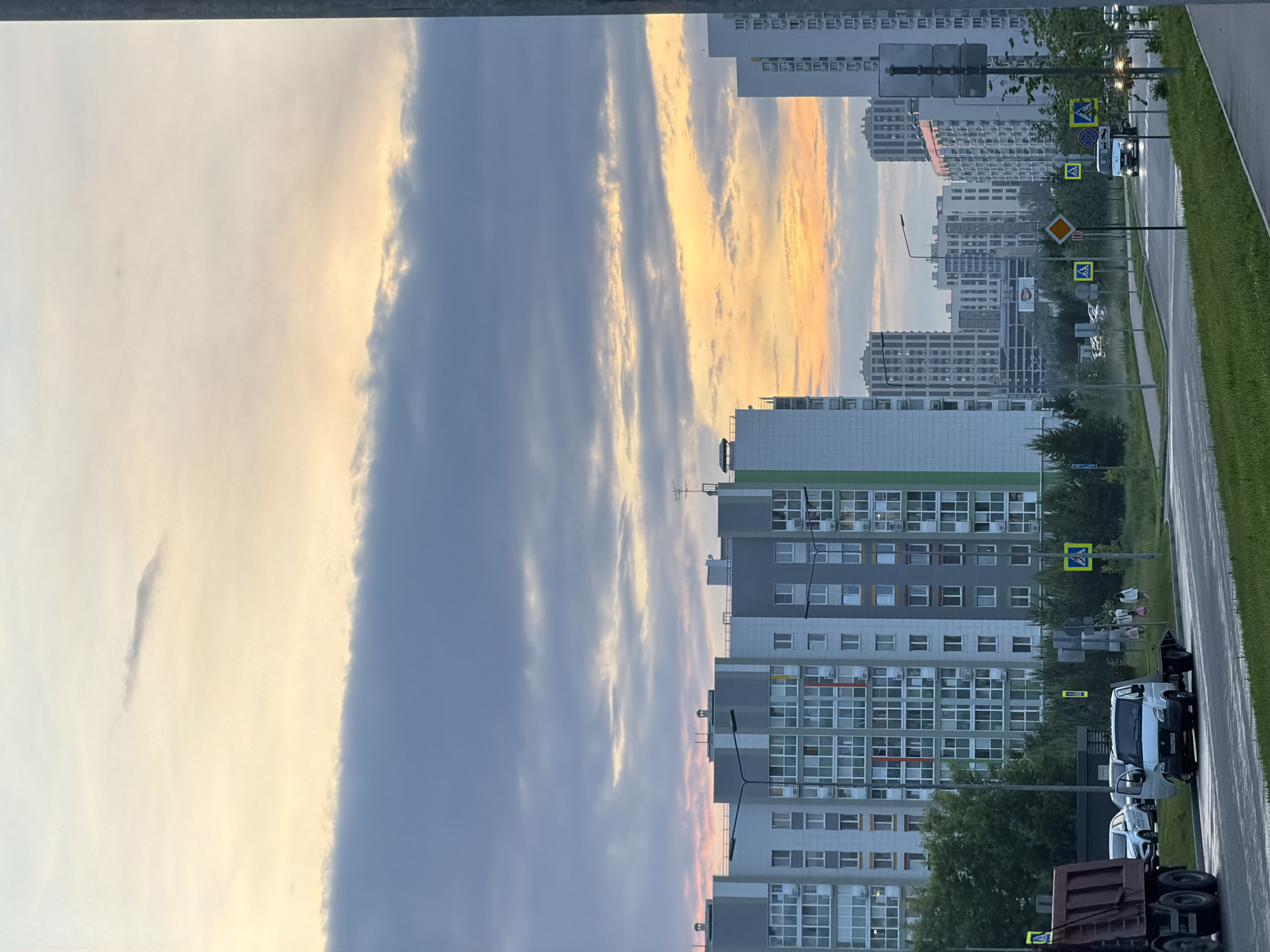 This is a medium-shot, eye-level, outdoor photograph taken in the city of Unknown, Unknown. The image depicts a street scene with apartment buildings and a cloudy sky. In the foreground, a wet asphalt road runs diagonally from the lower left to the upper right of the frame, bordered by a grassy median and sidewalk. A dark red dump truck is parked on the left side of the road, with its front facing right. To the right of the dump truck, a white utility van is parked, facing right. Further down the road, a dark-colored passenger vehicle is visible, heading towards the viewer. A series of pedestrian crossing signs with yellow backgrounds and black symbols are placed along the right side of the road, interspersed with streetlights and utility poles.
Behind the road, several modern apartment buildings of varying heights and architectural styles dominate the midground and background. These buildings are characterized by their grid-like window patterns, balconies, and a color palette of white, grey, and green. Trees and foliage line the street and surround the base of the buildings. The sky above is filled with a mix of dark grey and lighter, yellowish clouds, suggesting either early morning or late evening light. The overall impression is of a residential area under an overcast sky.