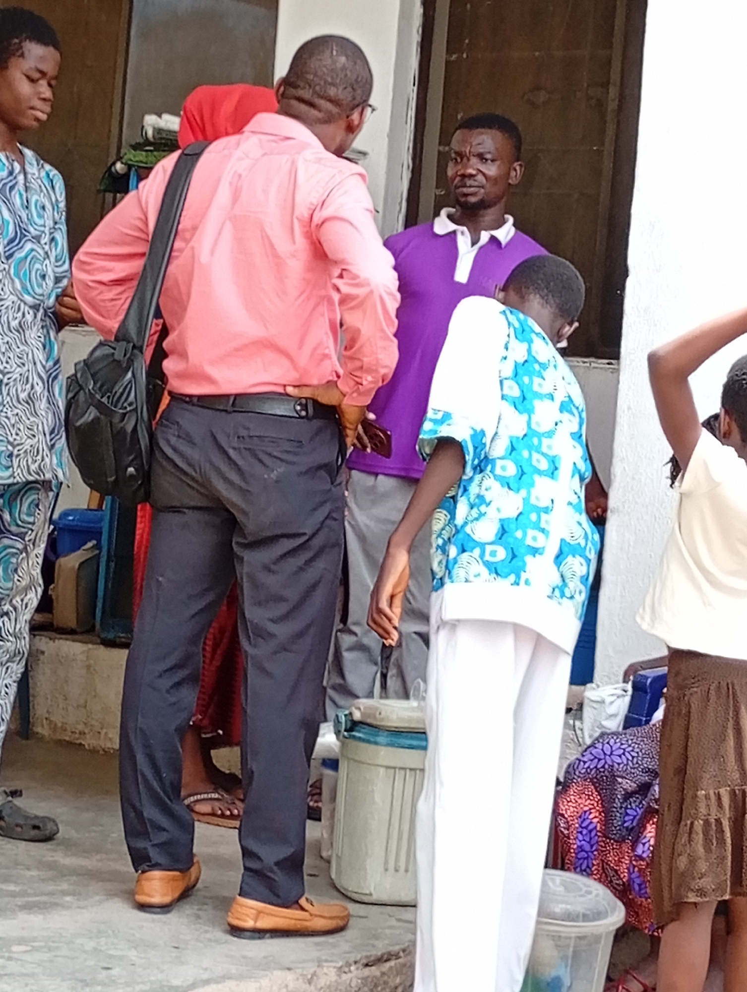 A group of people stands on a concrete surface, likely outdoors, near a building with a visible window. One man wears a pink shirt, dark pants, and tan shoes, with a black shoulder bag. Another man wears a purple polo shirt. A young person is dressed in a blue and white patterned shirt and white pants. Other people are partially visible in the frame, one wearing a blue patterned top, another a light-colored top, and a brown skirt. Various containers and belongings are scattered around the area. The location is identified as Lokoja, Nigeria.