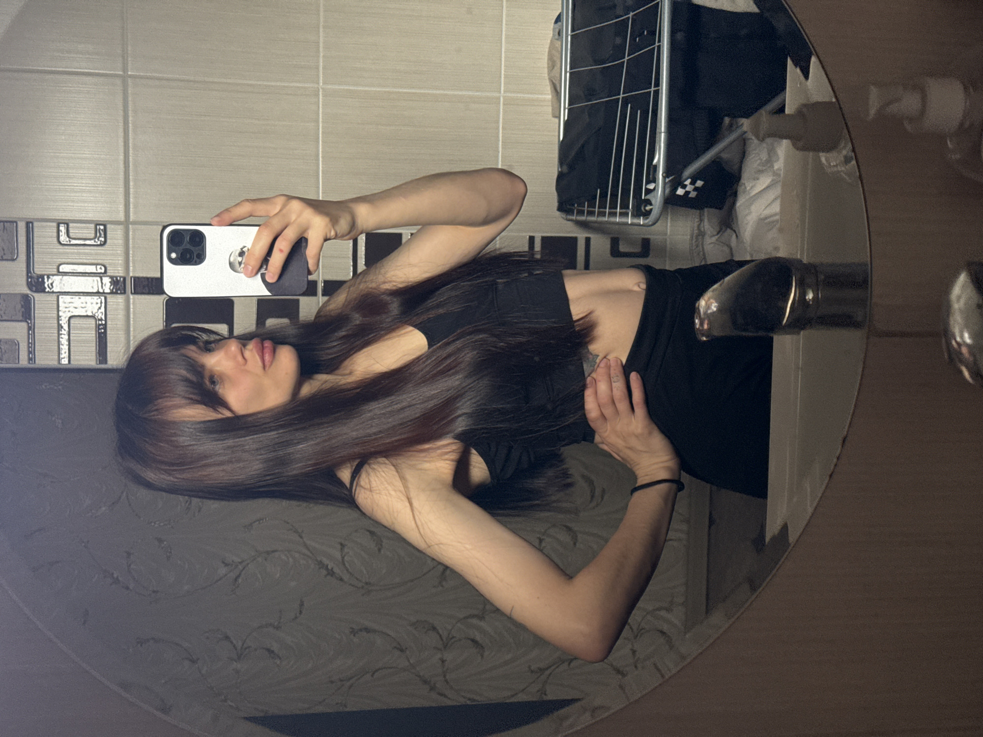 A woman is captured in a mirror selfie, holding a smartphone to take her picture. The image is rotated 90 degrees clockwise, but when reoriented, she is standing upright, looking into the phone's camera. She has long, dark brown hair with bangs that falls over her left shoulder and down her back. She is wearing a black sleeveless top. Her left hand holds a white or light-colored smartphone with a prominent black triple-lens camera module and a black pop socket. Her right hand is placed gently on her abdomen. She has a neutral expression on her face.
The setting appears to be a bathroom, as indicated by the light-colored rectangular wall tiles with some decorative dark geometric patterns. A silver-colored faucet is visible in the lower right portion of the mirror's reflection. Above the faucet, a metal shelving unit, possibly a shower caddy or storage rack, holds clothes or towels. Various toiletries, including bottles, are also reflected. Below the mirror's reflection of the woman, a dark, possibly patterned, surface is visible, which could be part of the vanity or floor. The mirror itself has a curved edge, suggesting it might be oval or irregularly shaped. The lighting appears to be artificial. No discernible text is visible in the image. This scene is located in Rostov-on-Don, Russia.