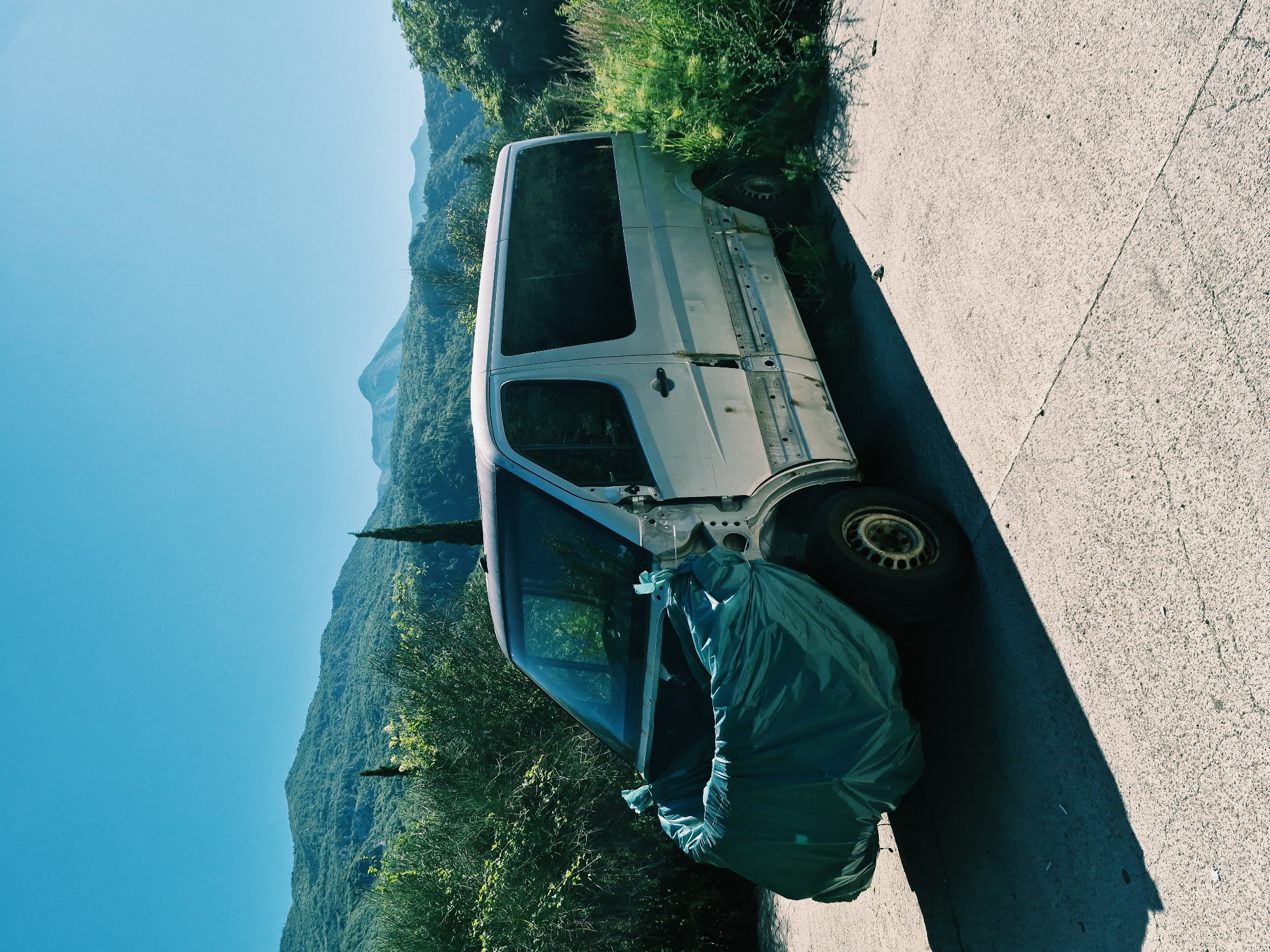 An abandoned, light-colored panel van, its front end covered by a teal tarp, is parked on a paved surface with a cracked texture. The van appears to be in a state of disrepair, with its front fender and possibly the hood missing, exposing a section of the engine bay. The driver's side of the van shows damage to the body panels below the windows. Green foliage, including tall grasses and bushes, grows around the vehicle, encroaching on its lower sections and the surrounding area. In the background, rolling, tree-covered hills rise under a clear, bright blue sky. The scene is lit by harsh sunlight, casting distinct shadows on the pavement. The location is identified as Prijevor, Montenegro. There are no people visible in the image.