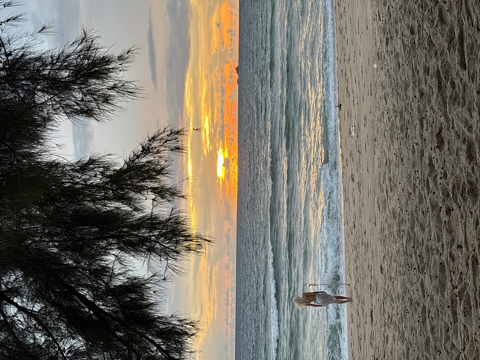 This is an eye-level, medium shot photograph taken at a beach in Choeng Thale, Thailand, during sunset. The foreground is a sandy beach with ripples and small dark objects scattered across it. In the mid-ground, a woman with blonde hair, wearing a light-colored, possibly sequined, dress, stands facing the ocean. She is positioned near the water's edge, and a tripod with a light or camera is next to her. The ocean is calm with gentle waves rolling onto the shore. The background features a vibrant sunset with hues of orange, yellow, and pink in the sky, with scattered clouds. A dark silhouette of a distant island is visible on the horizon. Overhanging branches of a tree frame the top of the image, adding a natural border to the scene.