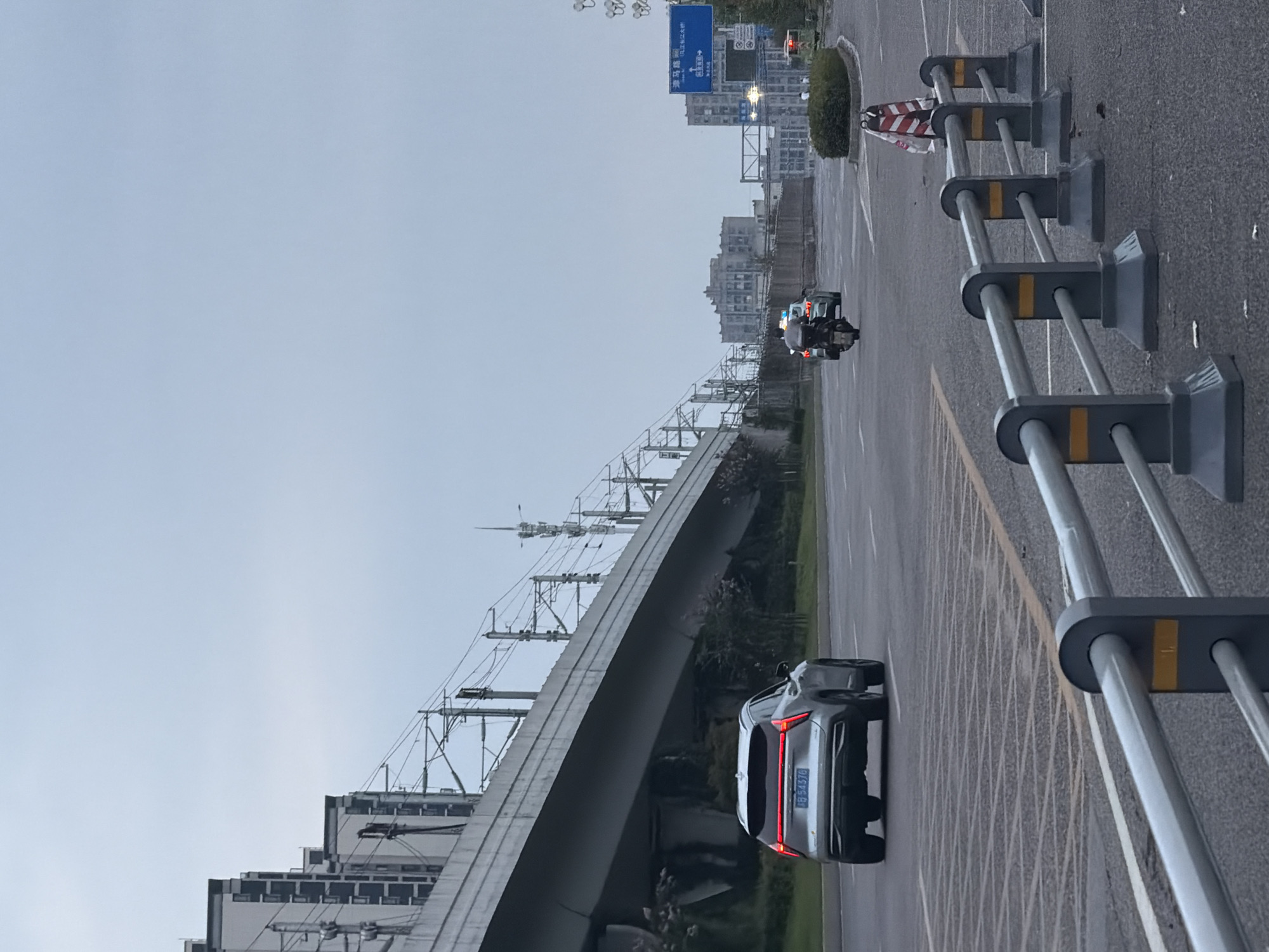 The image depicts an overcast day scene on a road in Jiangjin, China. In the foreground, a grey metal barrier runs along the side of the road. Beyond the barrier, a silver SUV with its taillights illuminated is driving away from the camera on a multi-lane road. Behind the SUV, a motorcycle is visible, also moving away. In the background, an elevated structure, likely a highway or railway overpass, dominates the left side of the frame. Further in the distance, buildings and a street sign are present. The sky is a uniform grey, indicating cloudy weather. There are no discernible people in the immediate vicinity of the vehicles or the barrier, though figures may be present in the background near the buildings. The road appears to be asphalt with lane markings.
