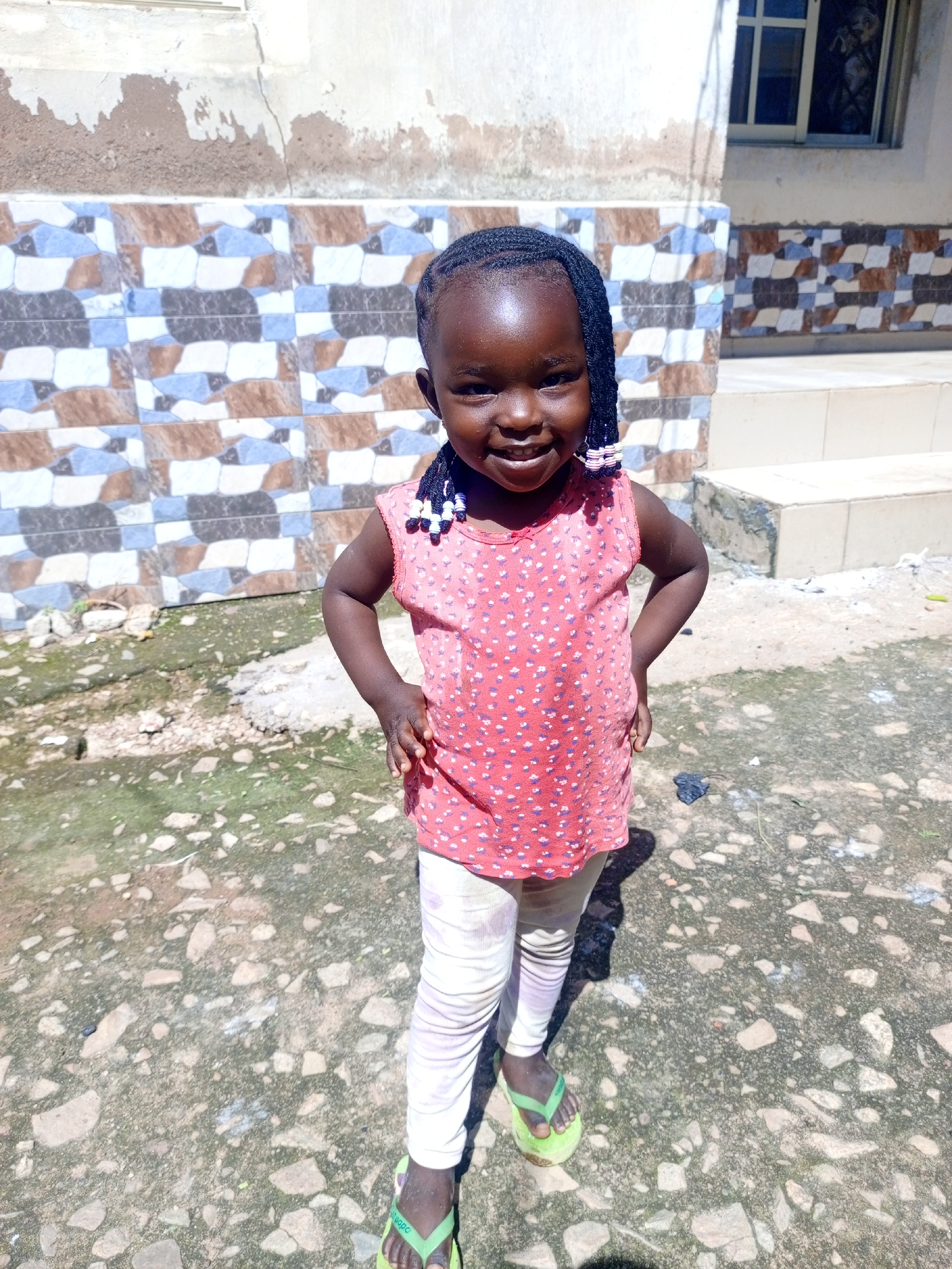 A young girl with dark skin and braided hair stands in an outdoor setting in Jos, Nigeria. She is wearing a pink sleeveless top with a white polka dot pattern and white leggings. Her hair is styled in braids with white and black beads. She is also wearing green flip-flops. The background features a tiled wall with geometric patterns in shades of brown, blue, and white, and a weathered wall above it. The ground is a mix of dirt, small stones, and some patchy green vegetation. A window with bars is visible on the right side of the building. The lighting suggests it is daytime.