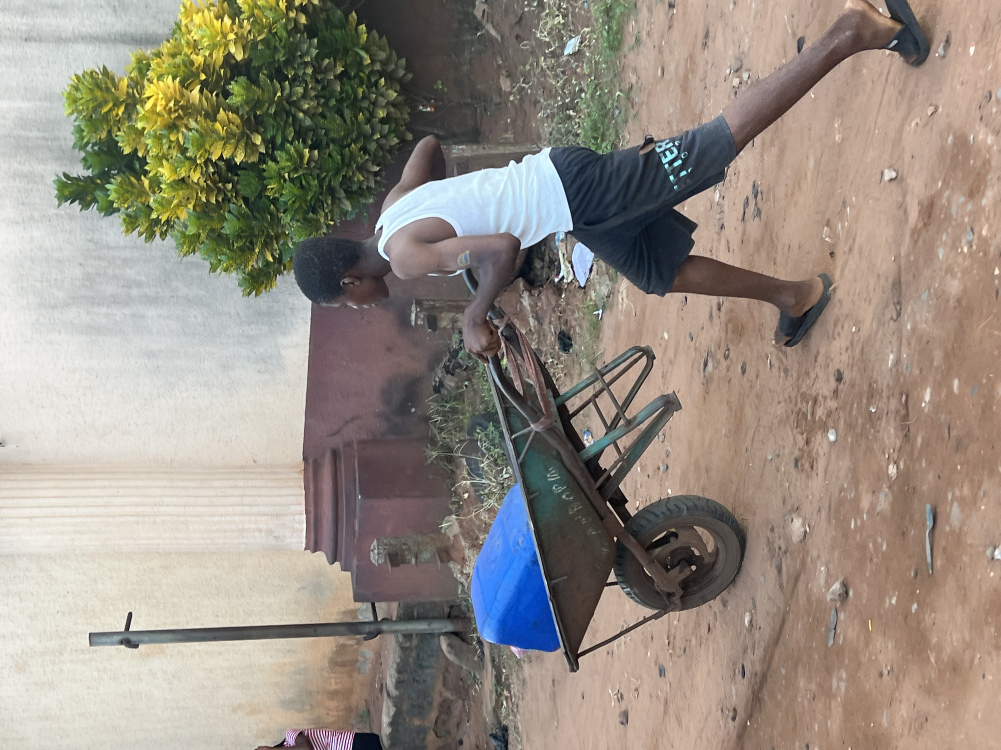 A young boy wearing a white tank top and black shorts with the word "BETTER" visible on the right leg pushes a wheelbarrow loaded with a blue plastic container. The boy is walking on a dirt path in what appears to be a residential area. Behind him, a building with a light-colored facade and a reddish-brown lower section is visible, along with a large, leafy green and yellow shrub. To the left of the boy, a person in a striped shirt is partially visible. The wheelbarrow is green and metallic, with a single wheel. The overall setting suggests a warm, outdoor environment. This scene is located in Nkpor, Nigeria.