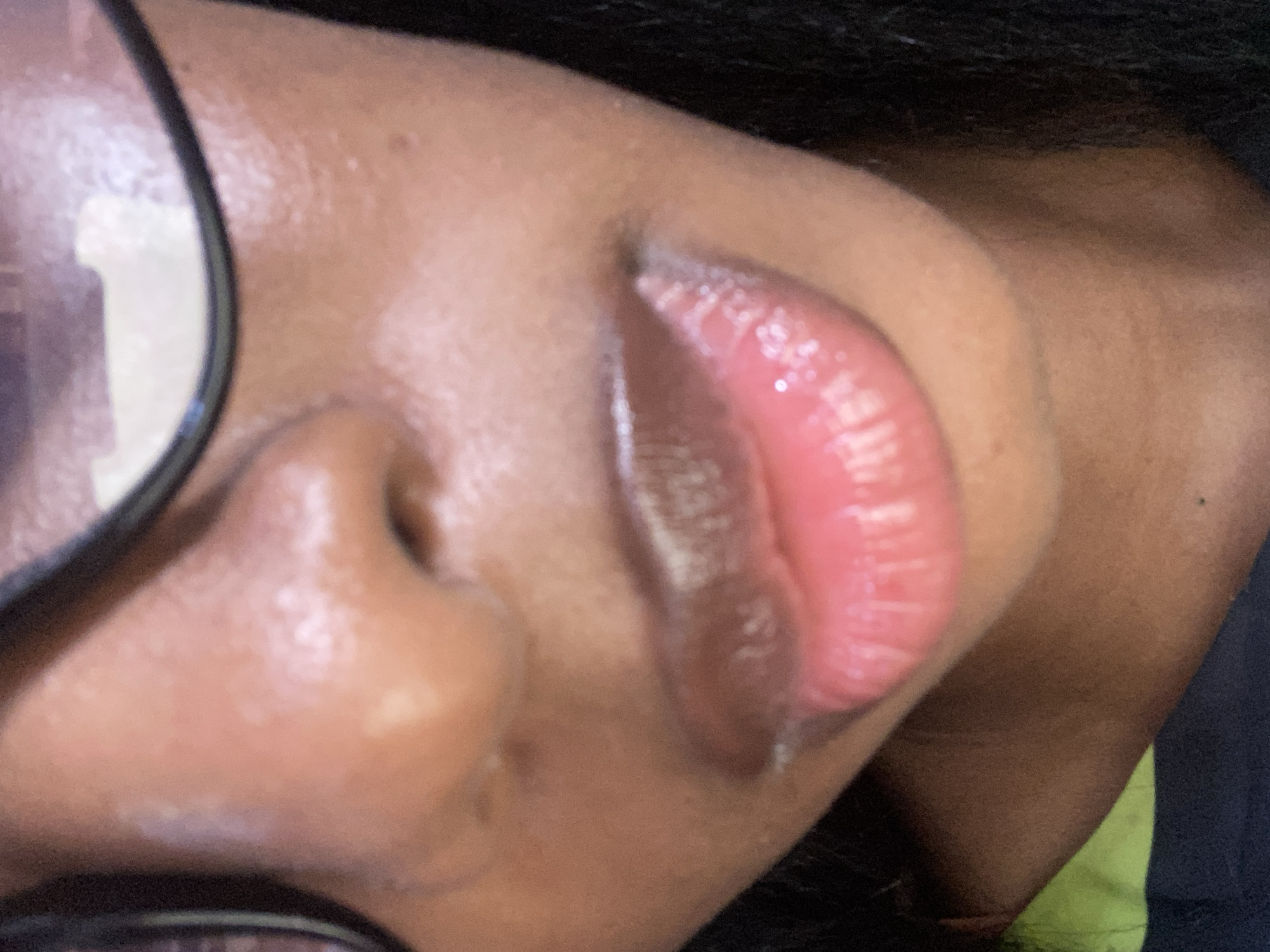 A close-up shot focuses on the lower portion of a person's face, specifically their nose and lips. The skin tone appears to be a medium to dark brown. The lips are slightly parted and have a glossy, pinkish-red appearance, suggesting the presence of lip gloss or balm. A pair of dark-rimmed glasses are partially visible at the top of the frame, obscuring the upper part of the face. The background is out of focus, but hints of dark fabric can be seen, possibly clothing. The lighting suggests an indoor setting or an area with soft, diffused light. The image is a portrait and does not depict any specific landmarks or elements that would indicate its location within Jos, Nigeria.
