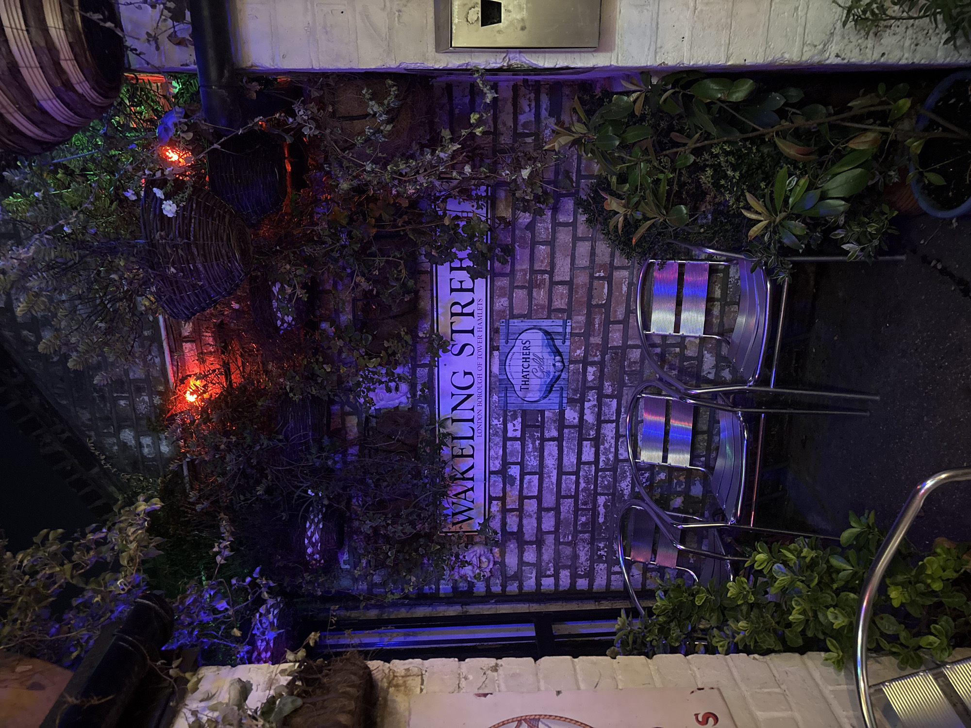 The image shows an outdoor scene, likely a patio or courtyard, with a brick wall as the backdrop. A street sign reading "Wakeling Street, London Borough of Tower Hamlets" is affixed to the wall, along with a sign that reads "Thatchers Gold." Wicker baskets containing plants hang on the wall. Two metal chairs sit near the wall, and additional plants and foliage surround the area. The scene is illuminated by various coloured lights, creating a dim, ambient atmosphere. The location is Wakeling Street in London, United Kingdom.