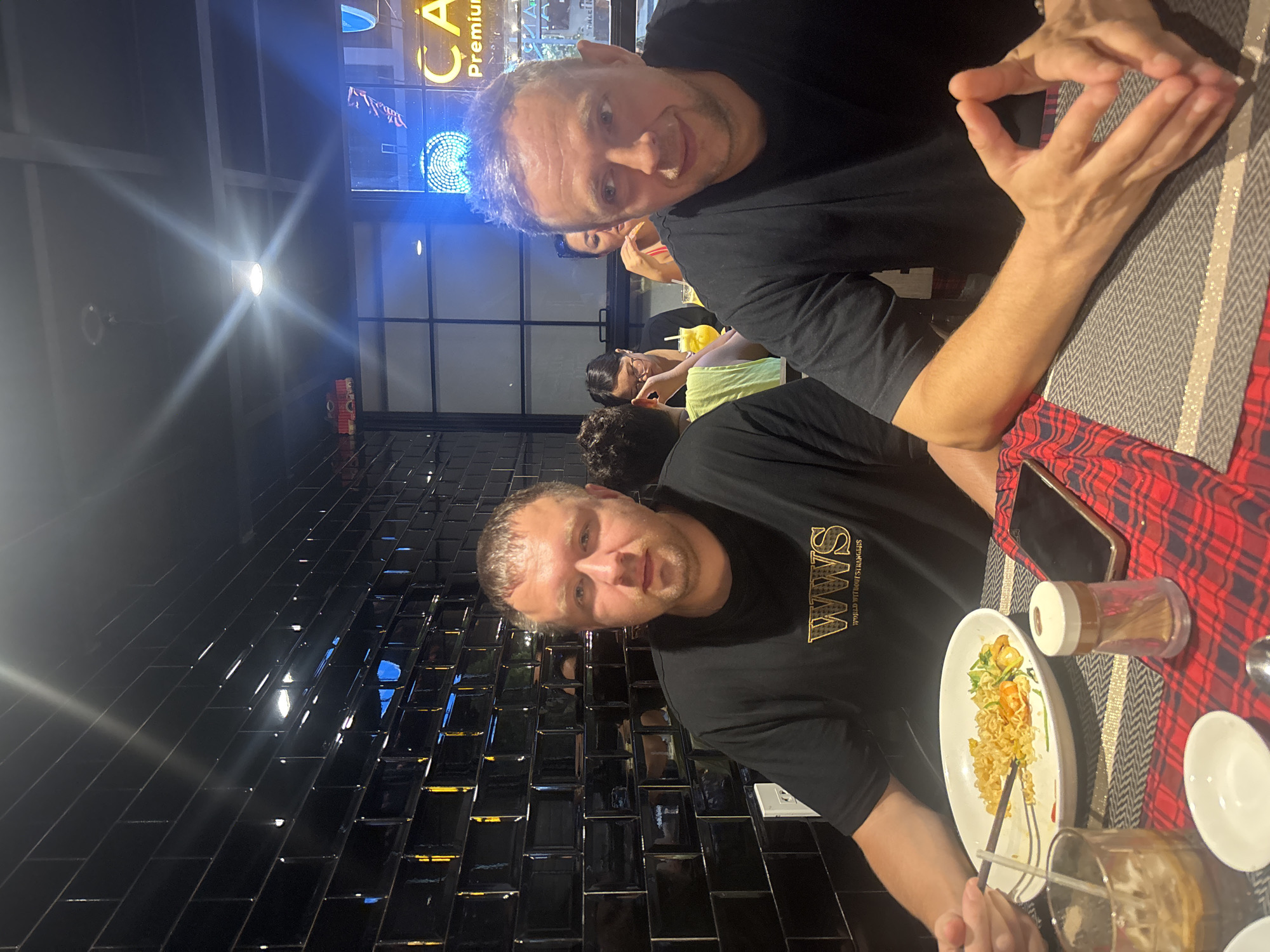 The image depicts an indoor scene within a restaurant or cafe, located in the city of Unknown, Unknown. Two adult males are seated at a table in the foreground, facing the camera.
The man on the left has light skin, short brown hair, and is wearing a black t-shirt with "WWS WORLD WITHOUT STRANGERS" in gold lettering. He is actively eating from a white oval plate containing noodles, shrimp, and green vegetables, using chopsticks in his right hand. A clear glass with a dark beverage and ice is positioned to his left, and a small white bowl is in front of him. A smartphone and a clear plastic toothpick holder are on the table to his right. The table is covered with a red and black plaid tablecloth over a gray placemat.
The man on the right, seated beside the first, also has light skin and short, graying hair. He wears a black t-shirt and is looking towards the right side of the frame with a slight smile. His hands are interlocked on a gray placemat.
The background on the left features a wall covered in dark, glossy subway-style tiles, reflecting ambient light. The background on the right reveals large glass windows or partitions. Through these, a bright blue neon light, forming a spiral or concentric circles, is visible. Above this, partial text "CA Premium" is discernible, likely part of an outdoor or interior sign. Further into the background, other patrons are seated at tables; one woman with dark hair and glasses is looking down, while another woman is partially visible behind her. Two yellow beverages are on their table. Overhead, a bright light fixture with a starburst effect is visible against a dark ceiling with visible structural framing. The overall lighting is artificial, casting reflections and creating bright points of light.