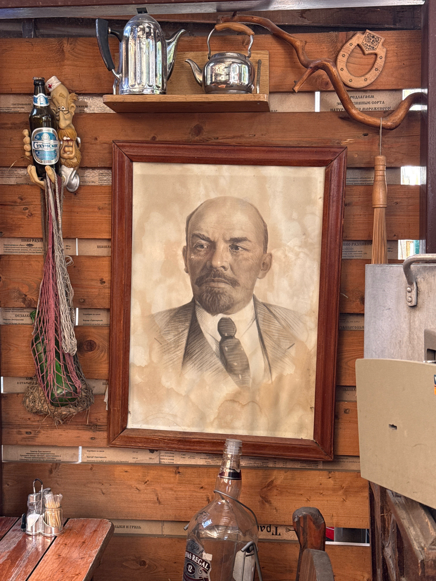 The image depicts an interior scene with wooden walls. A framed portrait of Vladimir Lenin is prominently displayed. Above the portrait, a shelf holds two metal teapots. To the left of the portrait, a bottle of "Okhumskoye" beer hangs from a decorative holder shaped like a person. Below the portrait, a large empty bottle of Chivas Regal is visible. In the foreground, a wooden table is set with salt and pepper shakers and a container of toothpicks. The environment appears to be a rustic establishment, possibly a cafe or bar, in Zvenigorod, Russia, given the context.