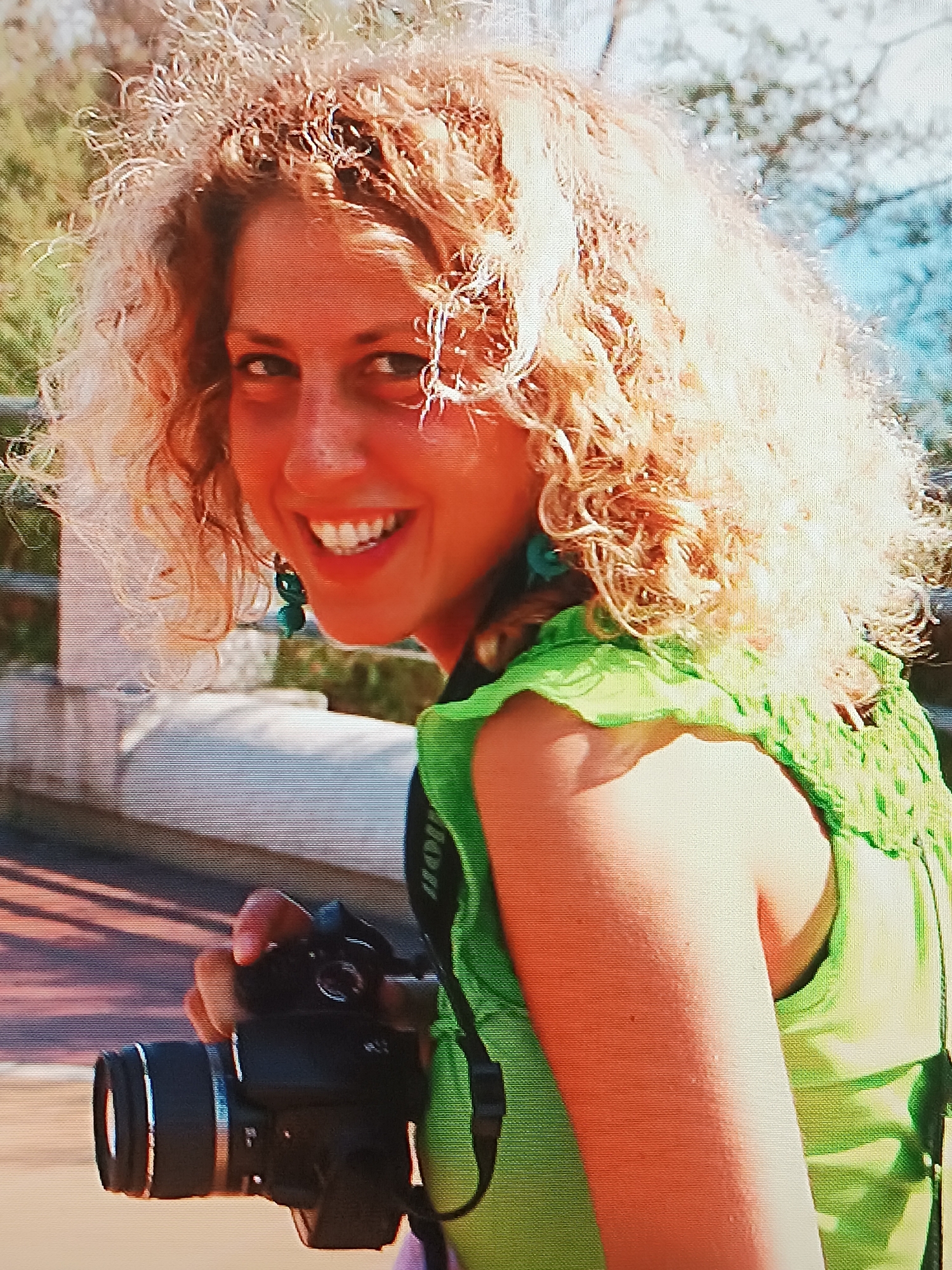 A woman with curly blonde hair and a bright smile holds a black camera. She is wearing a green top and turquoise earrings. The background suggests an outdoor location, possibly a bridge or elevated walkway, with foliage and a hint of sky visible. Although the city of Tbilisi, Georgia is mentioned in the prompt, there is no evidence in the image to confirm the location.