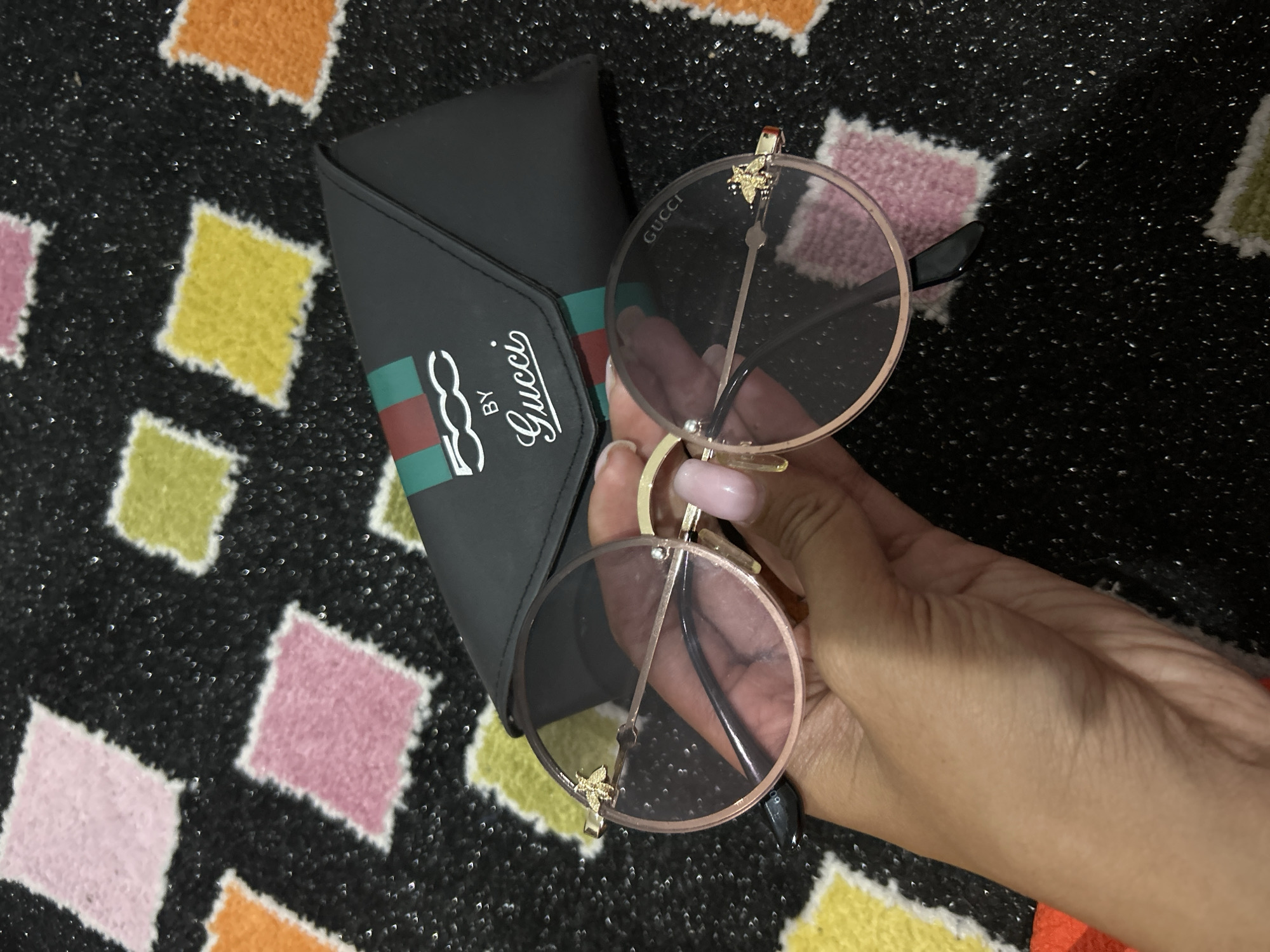 The image shows a pair of Gucci sunglasses, possibly aviator style, with rose gold frames and tinted lenses. A bee motif is visible on the left temple. The sunglasses are held by a person's hand with manicured nails. A black Gucci glasses case with the "500 BY GUCCI" logo is visible behind the sunglasses. The items are placed on a black background with a colorful diamond pattern. The location is not specified in the image or the prompt.