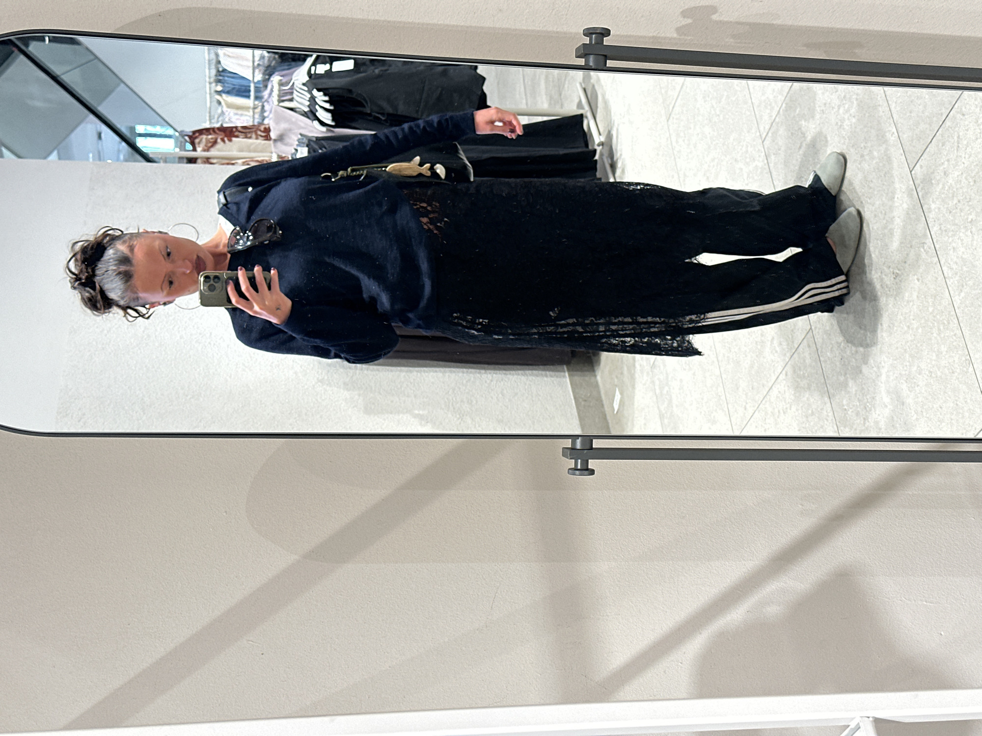 The image is a selfie taken in a mirror, likely inside a clothing store. The subject is a woman with her hair pulled up in a bun, wearing large hoop earrings and holding a smartphone. She is dressed in a dark sweater, a black lace dress, and black pants with white stripes down the side. Sunglasses are perched on her sweater.
The reflection in the mirror shows clothing racks in the background, suggesting the location is a retail environment. The floor is tiled with light-colored squares.
The location in London is unknown, as no landmarks or distinct architectural details are visible.