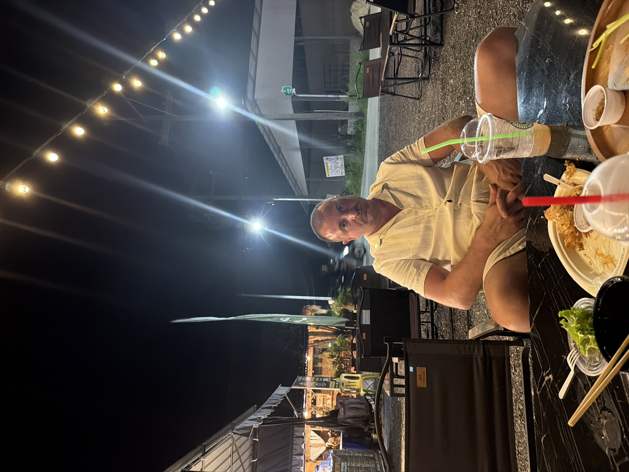 A man sits at a table outdoors at night, in what appears to be an open-air restaurant or market in Bang Muang, Thailand. The man is wearing a light-colored short-sleeved button-up shirt and shorts, and is looking directly at the camera. In front of him is a black marble table with a drink in a clear plastic cup with a green straw, chopsticks, a fork, a small bowl of lettuce, and some food on a white plate. In the background, there are string lights overhead, other tables and chairs, tents or market stalls, and some buildings. The ground is covered in gravel.