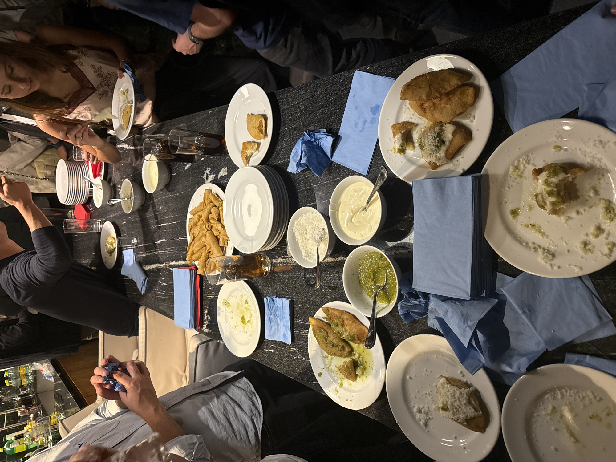 The image depicts a group of people gathered around a dark, possibly marble, table laden with food. The location appears to be in Mexico City, Mexico.
The table setting includes an assortment of white plates, some empty, others containing remnants of food, suggesting a meal in progress or recently completed. Fried food items, possibly empanadas or similar, are present on several plates. There are bowls filled with various condiments, including a white sauce, a green salsa, and what appears to be grated cheese. Stacks of blue napkins are scattered across the table. Glasses, some containing red wine, are also present.
Several people are partially visible around the table, engaging in conversation and presumably eating. One person is holding a blue napkin. The background suggests a somewhat dimly lit indoor environment, possibly a dining area in a restaurant or a private home. A bar area stocked with alcohol bottles is visible at the bottom left corner of the image.