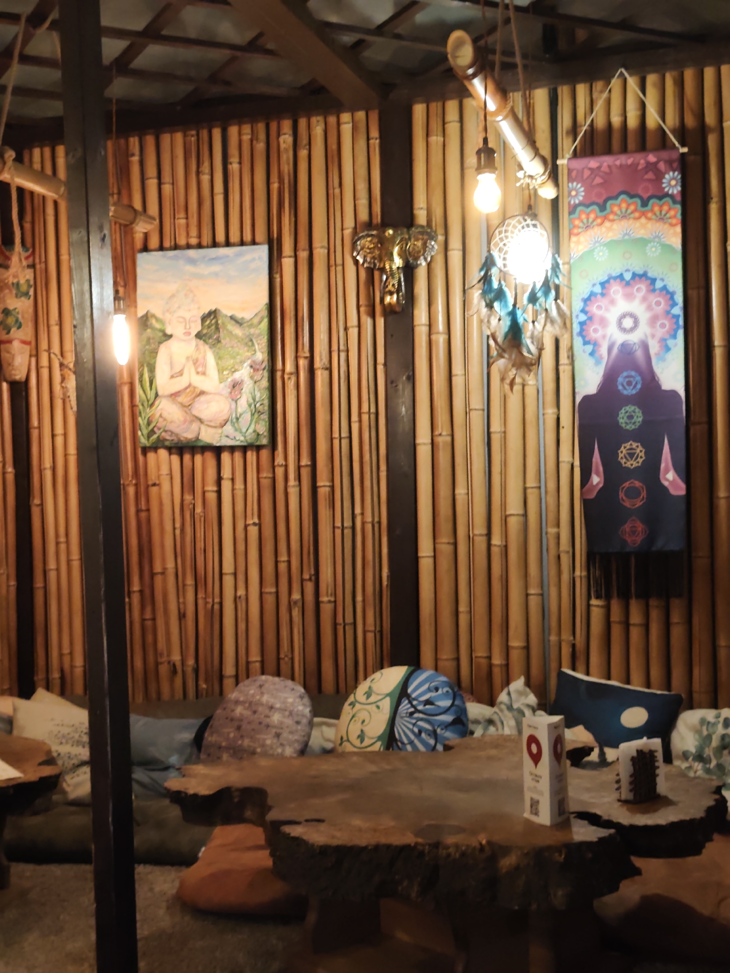 The image depicts the interior of a room with bamboo-paneled walls and a wooden ceiling. A painting of a meditating Buddha is on the left wall, with a decorative elephant head sculpture above it. To the right, a chakras tapestry hangs on the wall. In the foreground, a rustic wooden table sits in front of a seating area with cushions. A dreamcatcher hangs from the ceiling. The scene appears to be a relaxed, possibly meditation-themed space. No specific details suggest the location is Sochi, Russia.