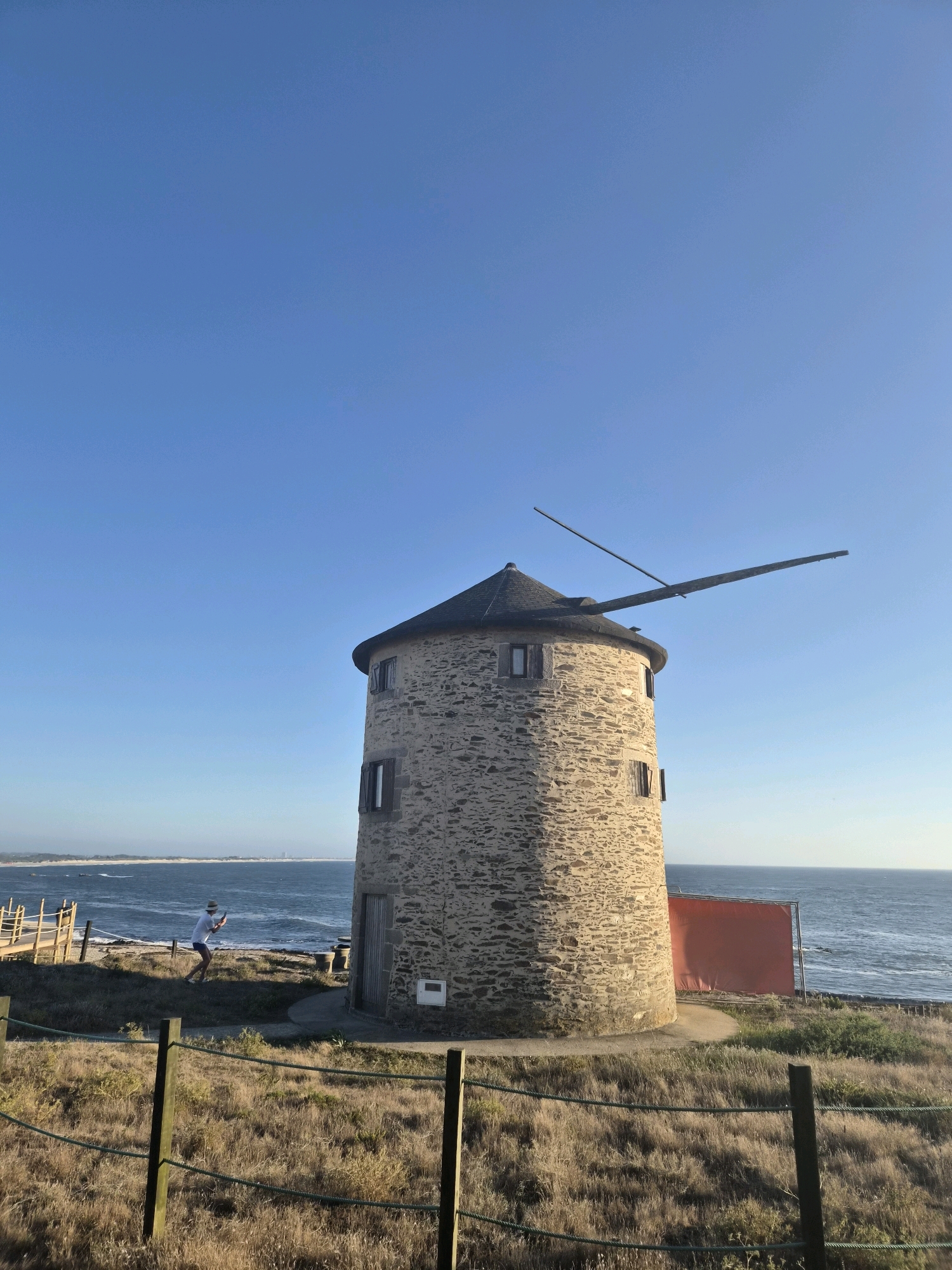 A stone windmill stands prominently on a grassy knoll overlooking the Atlantic Ocean, under a clear blue sky. The windmill, with its conical dark roof and two wooden blades, appears old and weathered. Small windows are visible on its cylindrical stone structure. A person wearing a white shirt and dark shorts, with a hat, is standing near the base of the windmill, facing away from the camera and looking towards the ocean, holding a phone, possibly taking a picture. The surrounding area is covered in dry, golden grass, with wooden fencing in the foreground. A bright orange tarp is visible behind the windmill, near the edge of the ocean. In the distance, a coastline with scattered buildings is visible. The scene suggests a peaceful, possibly tourist-oriented location, likely near the coast of Esposende, Portugal, given the architectural style and coastal setting.