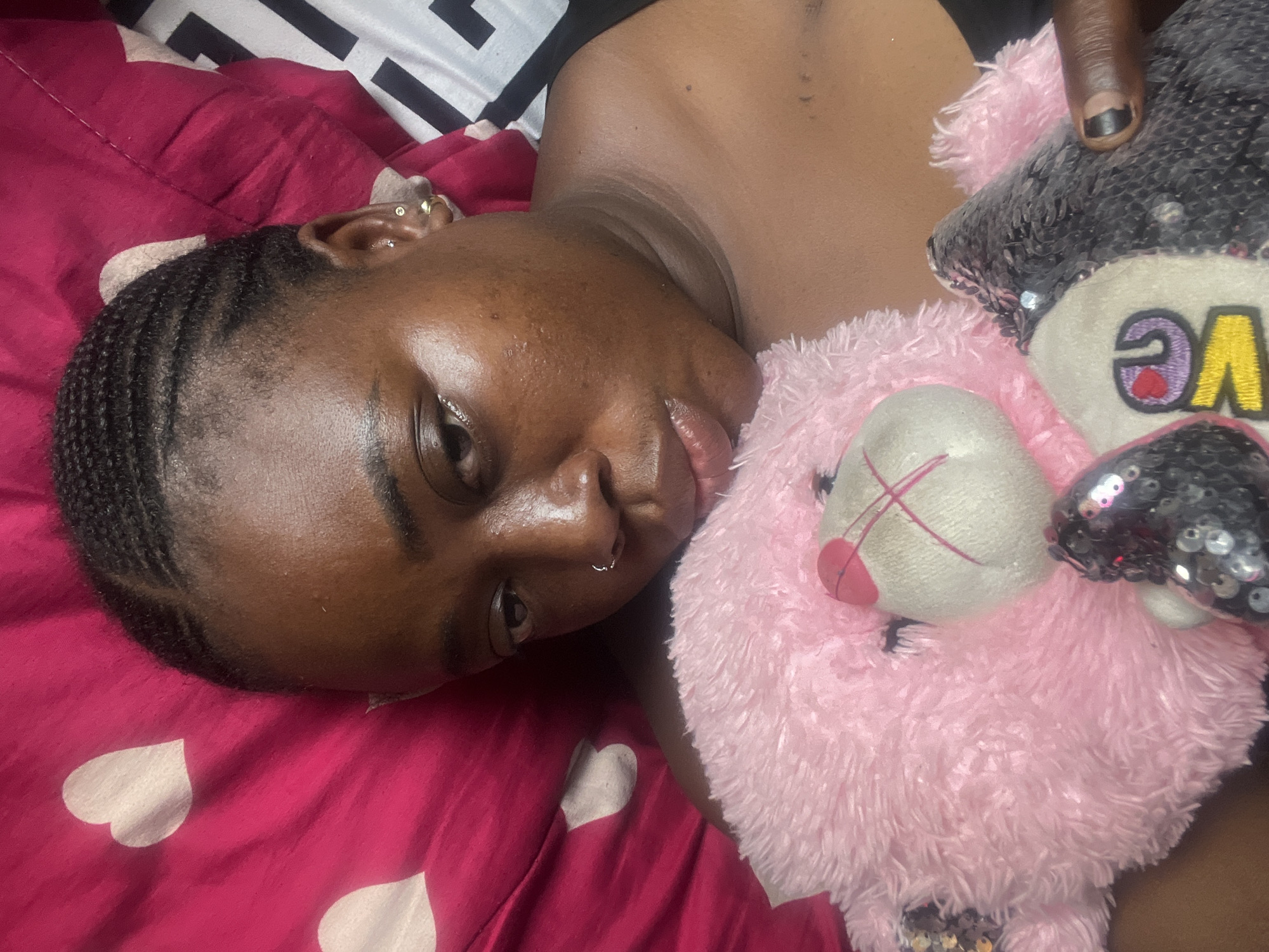 A young, dark-skinned woman with braided hair is lying on a pink and white heart-patterned pillow. She has piercings in her ear and nose. She is holding a pink, fluffy teddy bear with a white snout and crossed red lines for the mouth. The bear also has black sequin ears and "LOVE" embroidered in yellow with a heart. The woman's finger with black nail polish is visible near the bear. The background is a black and white patterned sheet. No information suggests the location is in Abuja, Nigeria.