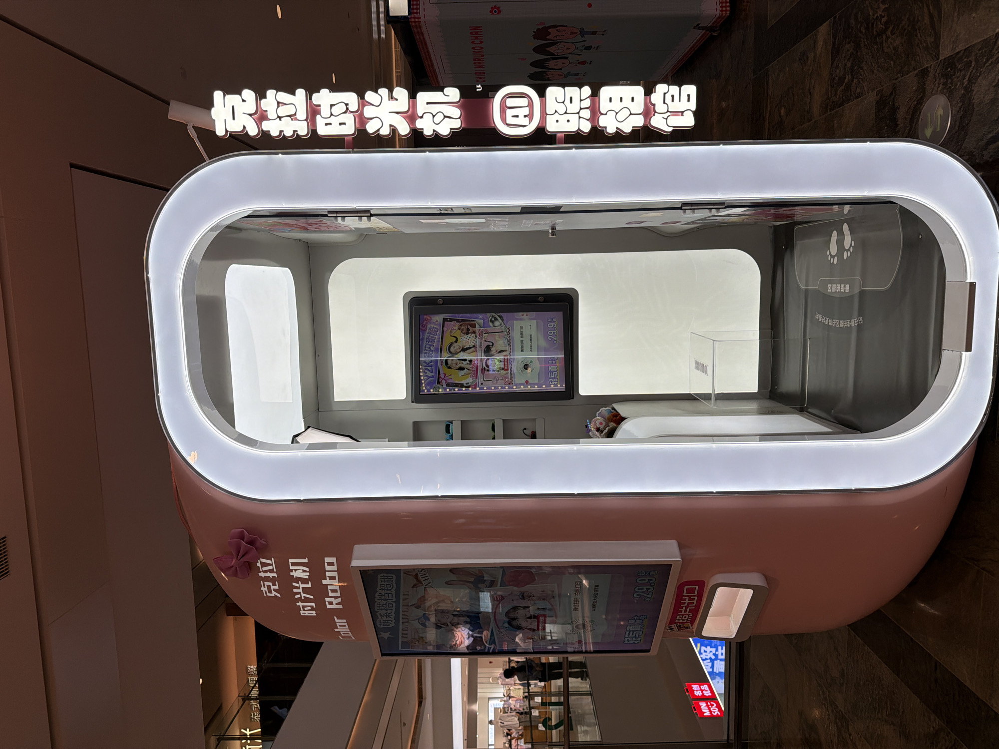 An illuminated, modern, pink and white self-service photo booth is centrally positioned in an indoor commercial setting, likely a shopping mall. The booth features a sleek, rounded design with bright white LED lighting framing its entrance.
Above the booth, a large, glowing sign reads "克拉时光机 AI 照相馆" (Color Robo AI Photo Studio). The exterior side of the booth displays "克拉 时光机 Color Robo" along with a screen showing vibrant, anime-style photo examples, prices like "29.9", and promotional text such as "萌系搭档超甜" (Cute Partner Super Sweet) and "2K银闪甜酷" (2K Silver Flash Sweet Cool). A small pink bow is affixed to the side. Below the display, a slot is labeled "照片出口" (Photo exit).
Inside the booth, a central screen offers various photo options and pricing, including "轻写真 29.9". Footprint decals on the floor indicate the "最佳拍摄区" (Best shooting area) and advise users to "站在最佳拍摄区拍得更好看的" (Stand in the best shooting area for better photos). Instructions like "微信扫码拍照打印" (WeChat scan to print photos) are also visible.
The background consists of dark, textured walls. Reflections on the booth's surfaces show parts of other storefronts, notably a "CHIBI MARUKO CHAN" sign above and reflections that suggest a "MINISO" store ("MINI 名创 优品") nearby. The setting appears well-lit, characteristic of a retail environment. The booth is clearly designed for customers to take fun, themed pictures.