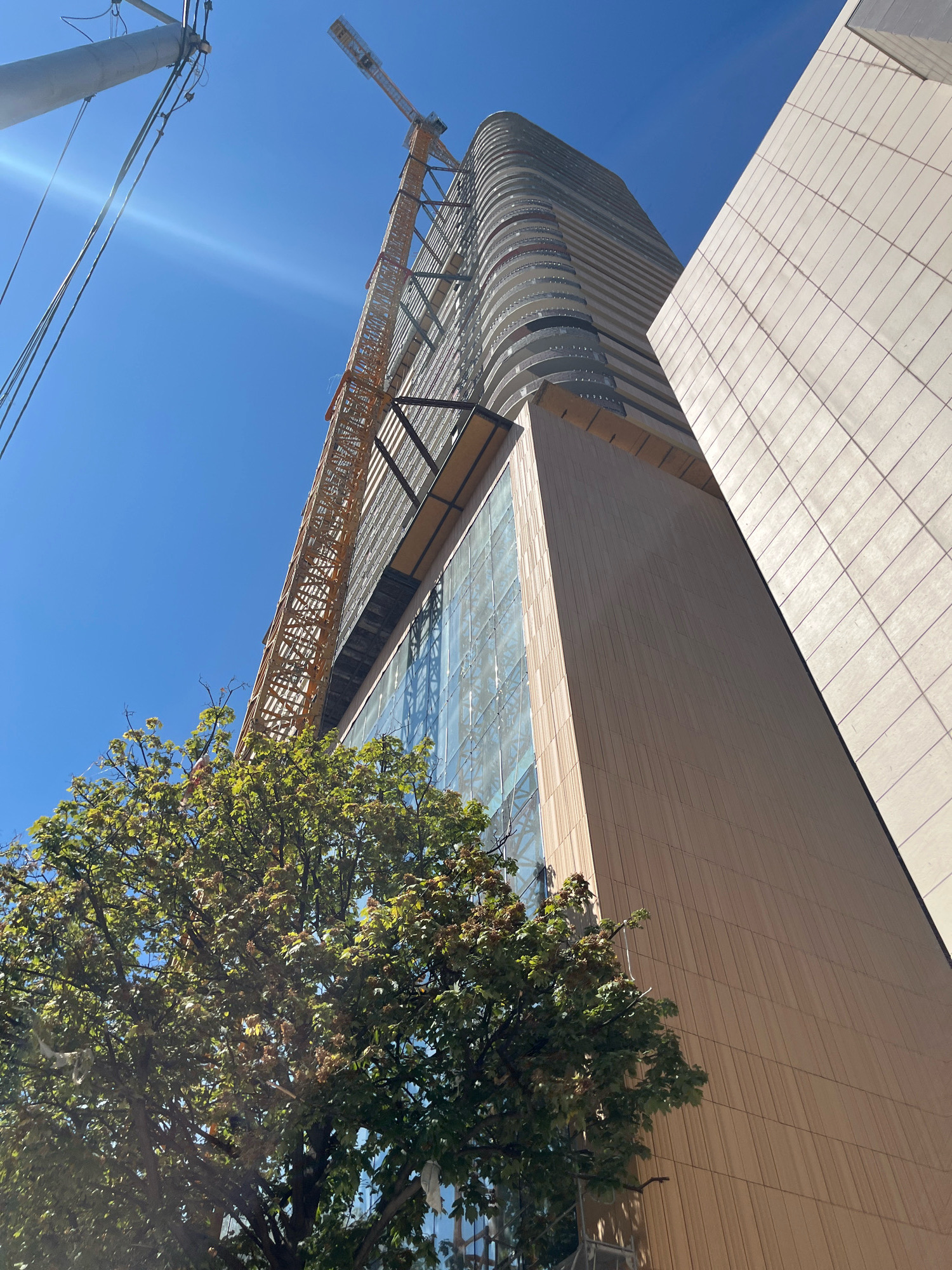 The image shows a construction site in Tbilisi, Georgia, with a tall building under construction and a yellow crane. The building has a modern design with curved balconies and glass windows. A smaller building with a tiled facade is adjacent to the taller structure. In the foreground, a green tree with lush foliage is partially visible. The sky is clear and blue. Power lines and a utility pole are present in the upper left corner of the frame.