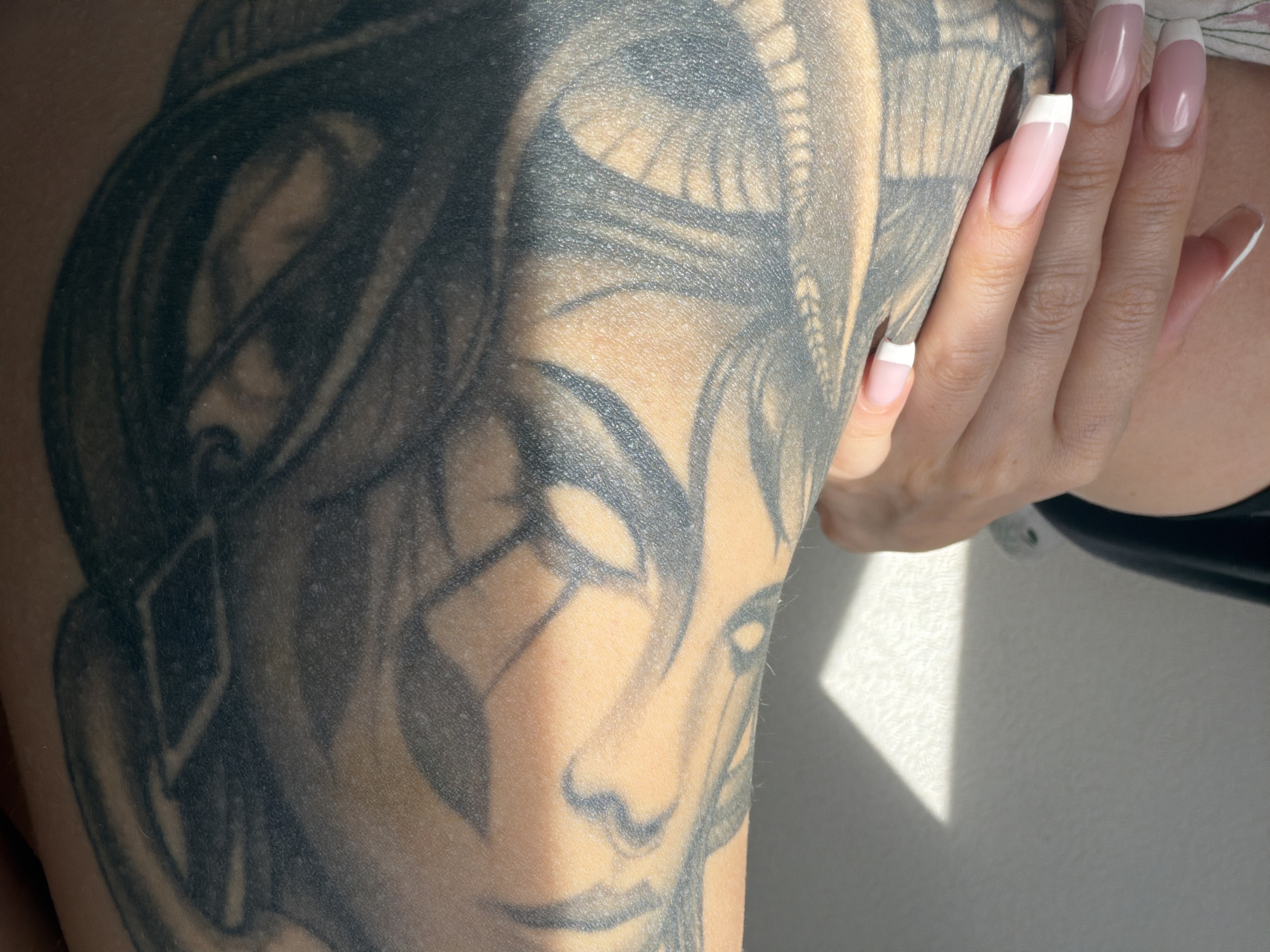 A close-up shot shows a person's arm with a large, black and gray tattoo featuring a face. The person's hand, with long French-manicured nails, rests on the arm. A white wall is visible in the background, partially lit by sunlight. The image provides no indicators of location in Saint Petersburg, Russia.