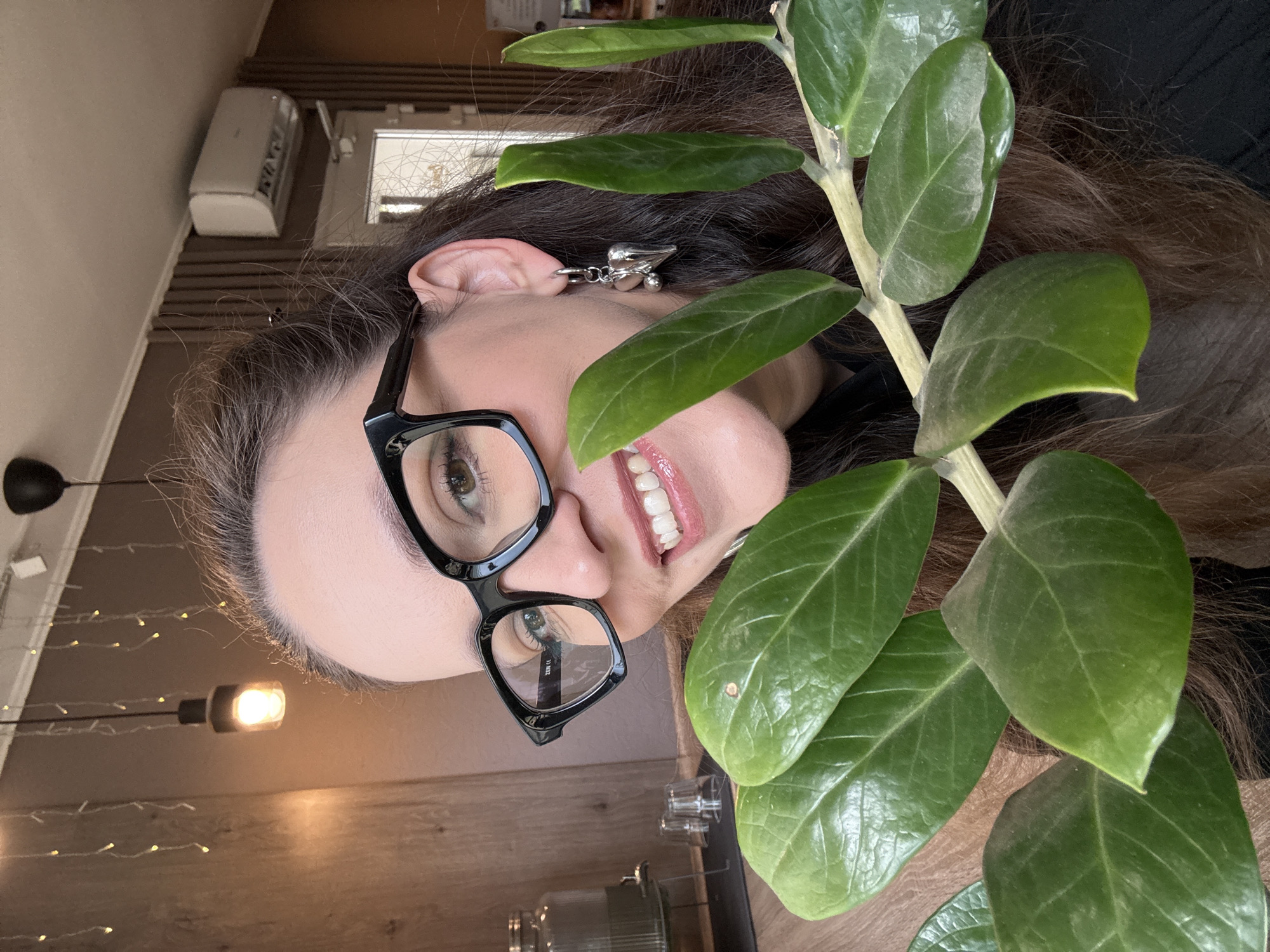 The image shows a close-up, medium shot of a woman with long, dark brown hair wearing black-rimmed glasses and a silver heart-shaped earring. She is smiling and looking towards the camera, peeking out from behind a branch with several large, green leaves. The background is an indoor environment with wood-paneled walls, decorative string lights, a ceiling light fixture, and an air conditioning unit mounted high on the wall. A glass beverage dispenser is visible on a shelf to the left. The lighting is warm and ambient. There is no discernible information in the image to suggest a specific location within the city of Chaikovsky, Russia.