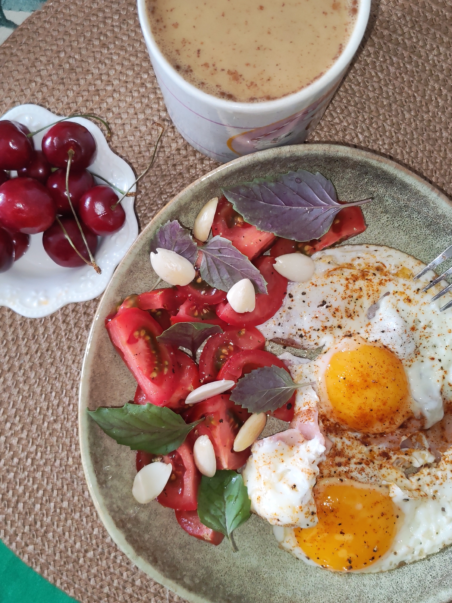 The image displays a top-down view of a meal arranged on a light brown woven placemat. A rustic-style plate, with a light green rim and textured beige-grey interior, occupies the central and lower-right portion. On this plate, two fried eggs are visible, topped with black pepper and a reddish-brown seasoning. A silver fork is positioned on the upper right egg. To the left of the eggs, sliced red tomatoes are intermingled with several whole, blanched almonds and both green and purple basil leaves.
In the upper left, a small, white, scalloped-edge dish contains a cluster of red cherries with their stems. Partially visible in the upper right, a white ceramic mug holds a light brown beverage with a frothy top, speckled with a darker powder. A sliver of a vibrant green fabric is visible in the bottom left corner of the frame, beneath the placemat.