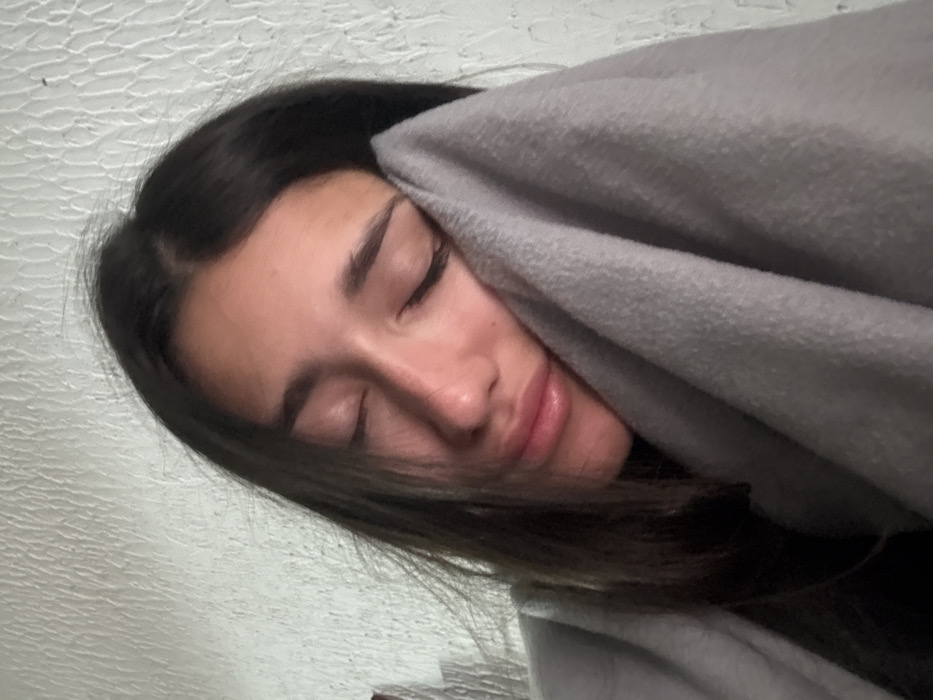 The image displays a close-up of a single individual, identified as a young adult female, resting in Zankovići, Montenegro. Her eyes are closed, and her head is tilted slightly to her right. She possesses long, dark hair. Her facial features include full, slightly parted lips. A light grey, soft-textured fabric, consistent with a blanket or shawl, is draped around her, partially covering her right cheek and chin. The background consists of a textured, light-colored wall with an uneven surface. The scene is illuminated by diffused, even lighting.