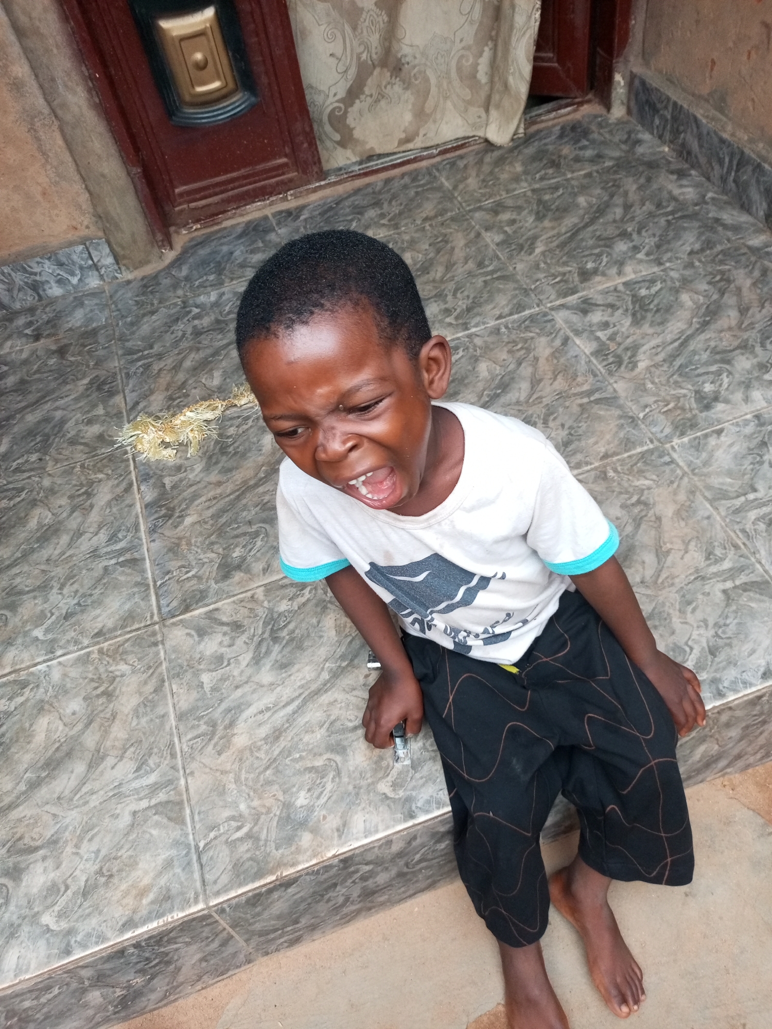A young Black boy is sitting on tiled steps outside, crying with his mouth open and eyes squeezed shut. He is wearing a white t-shirt with a blue trim and black pants with a brown wavy pattern. To his left on the steps is a small pile of dried, straw-like material. Behind him is a dark red door with a gold embellishment. The overall setting appears to be a residential area, possibly in a city like Tempure, Nigeria, characterized by its tiled flooring and simple building materials.