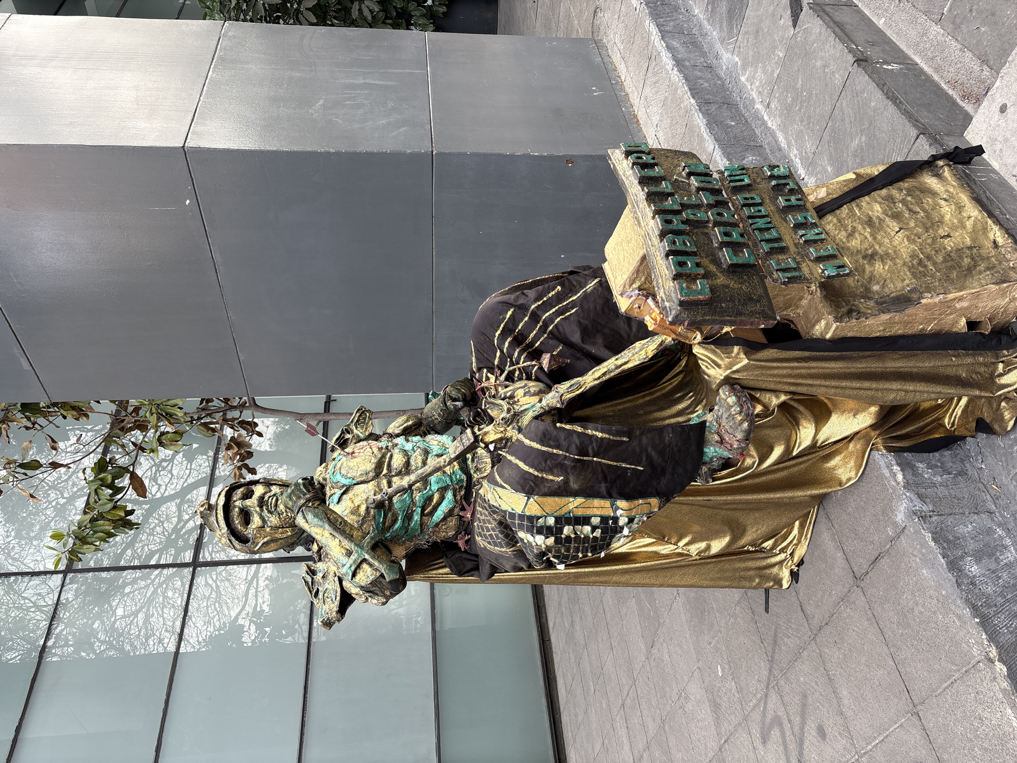 The image captures a street performer in Mexico City, Mexico, posing as a living statue. The performer is adorned in an elaborate costume of gold, black, and turquoise colors, designed to resemble an ancient warrior or deity. A sign, partially visible, is positioned next to the performer. The performer is located on a sidewalk adjacent to a modern building with a gray exterior and glass windows reflecting trees and sky. The setting is urban, with stone steps near the performance area. The overall scene suggests a common street entertainment display in a tourist location.