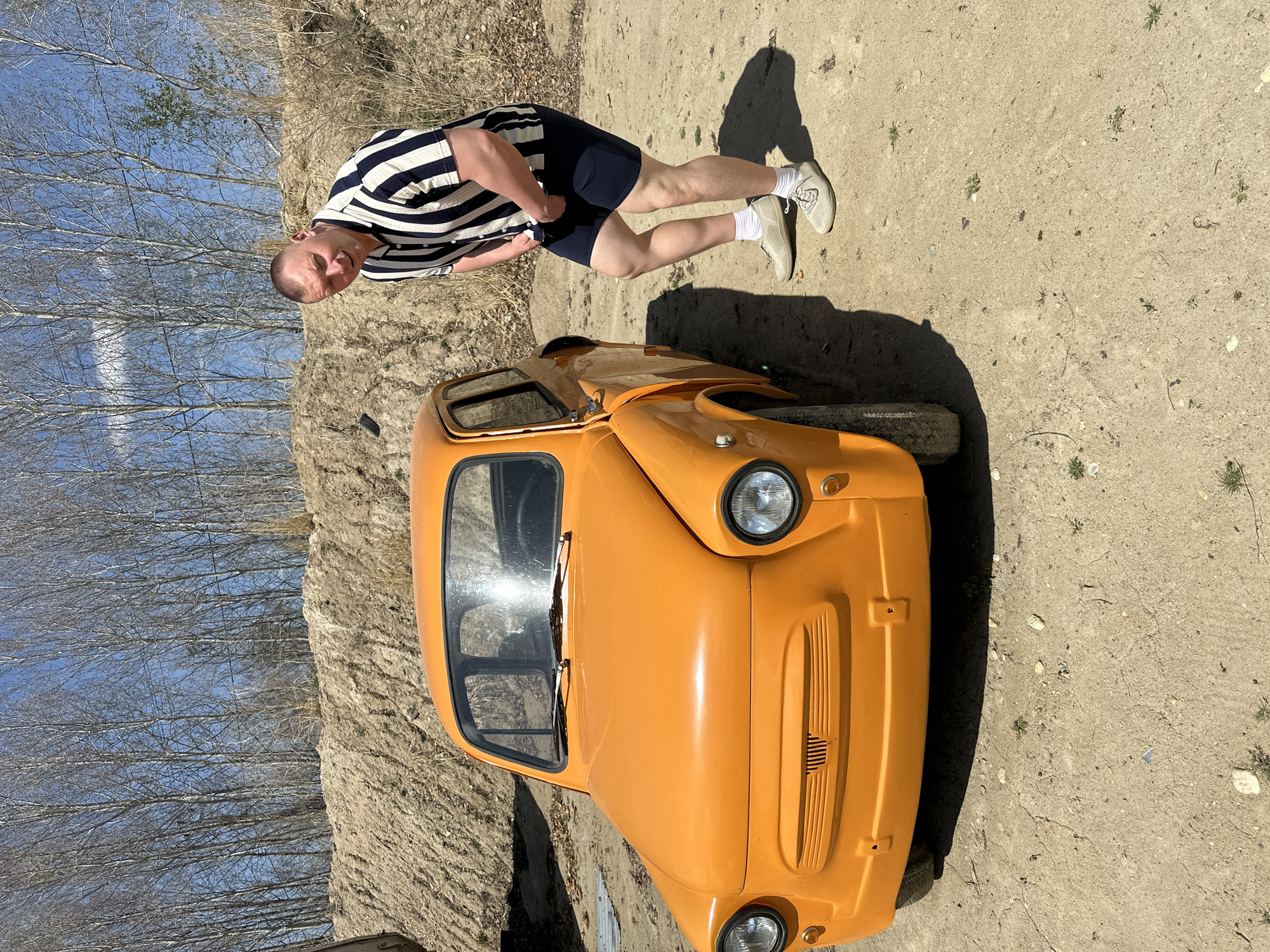 The image depicts a man standing next to a small, orange car. The man is wearing a striped shirt and navy shorts. The car is parked on a dirt surface with sparse vegetation. In the background, there are trees and a blue sky. The image was taken outdoors in Sadovoe, Russia.