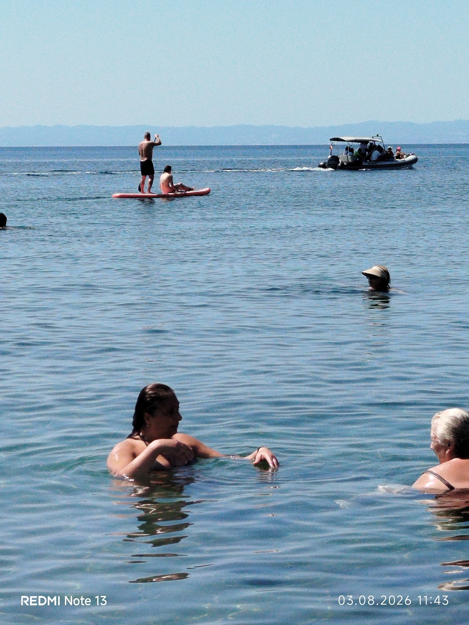 In the clear blue waters of Sithonia Municipal Unit, Greece, several individuals are enjoying a sunny day. In the foreground, two women are swimming, with one partially submerged and looking to her right, her arm raised. Another woman is further out, wearing a sun visor and looking to her left. In the mid-ground, a man stands on a paddleboard with another person seated behind him. Further in the distance, a boat with several passengers is moving across the water, leaving a wake behind it. The horizon is marked by a distant coastline with a clear sky above. The timestamp in the lower right corner indicates the image was taken on August 3, 2026, at 11:43 AM. The text "REDMI Note 13" is visible in the lower left corner.