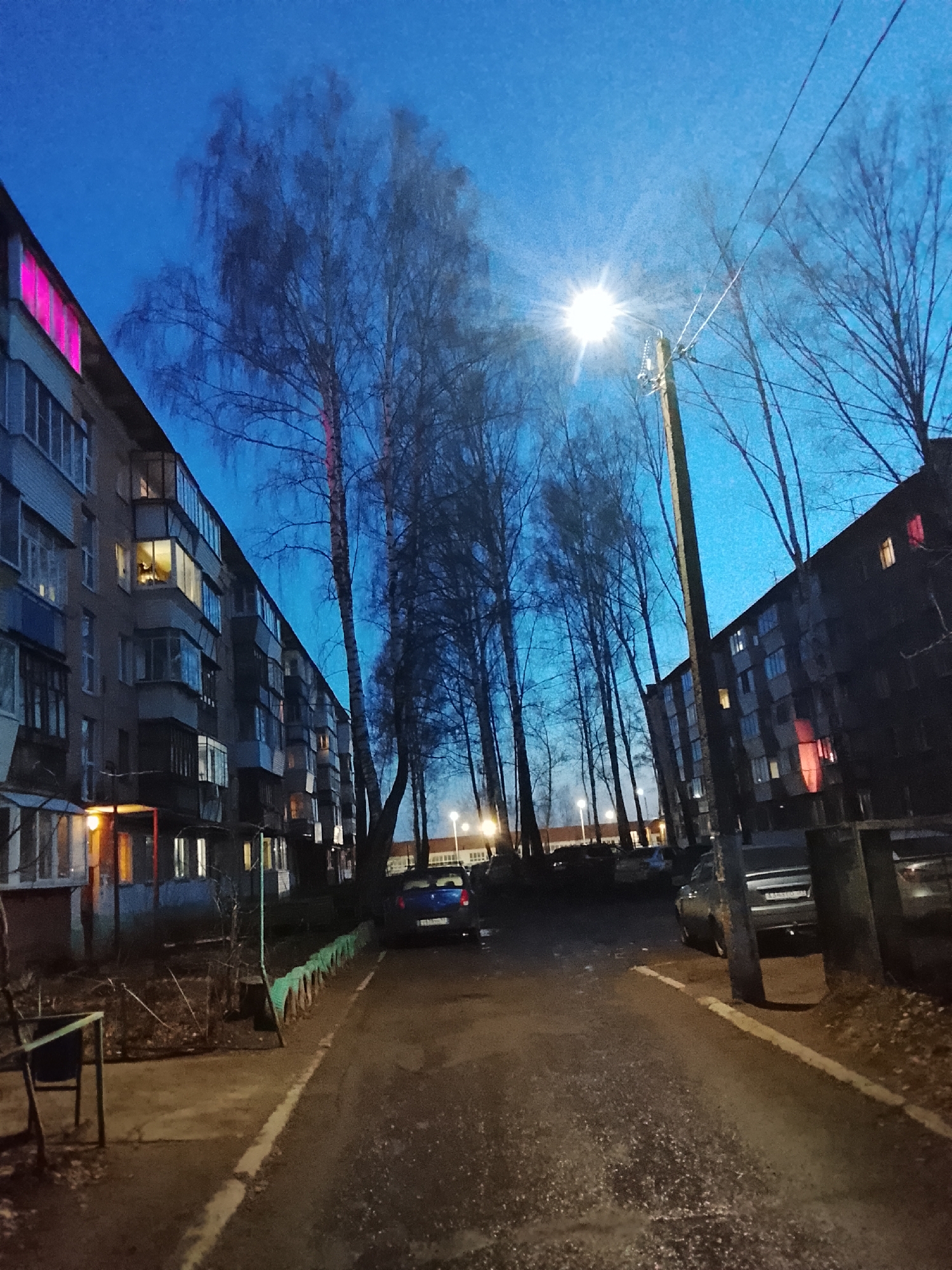 This image captures a quiet, residential street or courtyard in Chaikovsky, Russia, at dusk or early night. The sky is a deep, rich blue, characteristic of twilight hours, with artificial lights illuminating the scene.
Two multi-story apartment buildings, typical of Soviet-era architecture, flank a central paved path. The building on the left features numerous balconies, some enclosed with windows, and several windows show warm yellow light from within, suggesting occupancy. A distinctive, bright pink or magenta light emanates from a window on the upper floor of this building, adding a vibrant focal point. The building on the right is darker, partially obscured by trees and a utility pole, but some windows also emit warm light, and a bright red light is visible in one of its lower windows.
A prominent street light with a strong starburst effect casts a bright glow near the center-right of the image, illuminating the bare branches of tall deciduous trees that line the path. These trees, likely birches, stand silhouetted against the blue sky, their intricate branch patterns visible. A wooden utility pole with power lines is positioned directly beneath the brightest street light.
The ground consists of a damp-looking paved surface, possibly a road or courtyard, with a narrow curb running along the left side. A low fence, constructed from what appear to be repurposed green tires, borders a small patch of sparse vegetation near the left building. Several cars are parked along the sides, including a dark blue sedan visible on the left and another darker vehicle on the right. A partially visible Russian license plate on the darker car, showing characters such as "A340...", further indicates the location.
The scene conveys a calm, everyday urban atmosphere as residents settle in for the evening. No people are visible, contributing to the quietude of the residential setting. The cool blue tones of the sky contrast with the warm interior lights, creating a serene and somewhat contemplative mood.