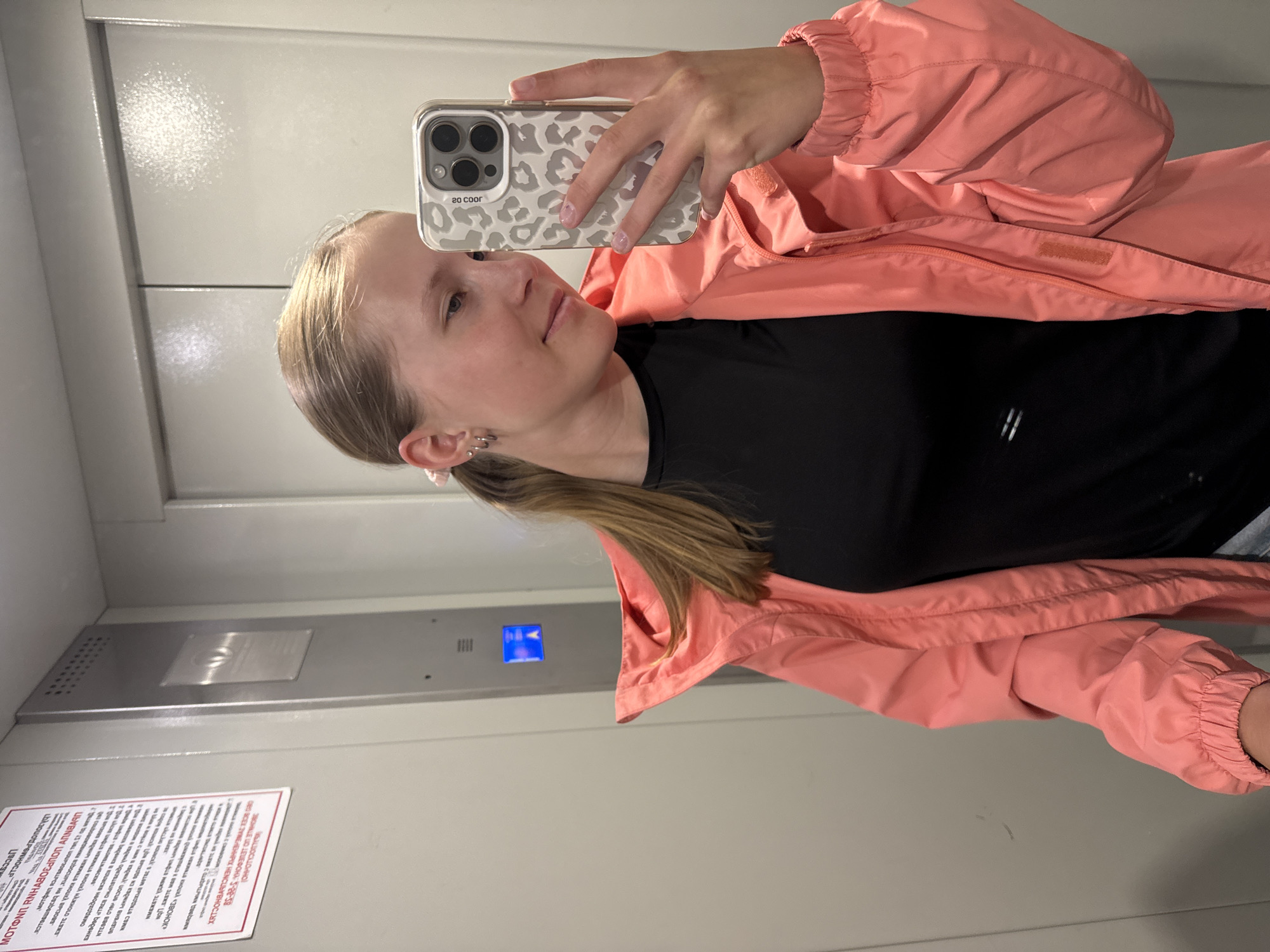 A young woman is captured in a mirror selfie inside an elevator. She is positioned facing slightly to her left, holding a smartphone with a grey leopard print case in her right hand, which reflects her image. Her light-colored hair is pulled back, and she wears a black top under a coral/pink lightweight jacket with elasticized cuffs. Small earrings are visible on her earlobes.
The setting is an elevator interior with light-colored, possibly reflective, walls. An elevator control panel with a blue illuminated button is visible on the wall to the left of the woman's reflection. Below this panel, a white rectangular sign with extensive text in Russian is affixed. The text, partially readable, includes phrases like "ЗВОНИТЕ ПО ТЕЛЕФОНУ" (Call by phone), "ВНИМАНИЯ" (Attention), and "МОТОРИН", suggesting it's an informational or emergency notice for the elevator's operation or repair. The phone case also displays "20 COOR" in print. The woman maintains a neutral, calm expression. The location context for the scene is Chaikovsky, Russia.