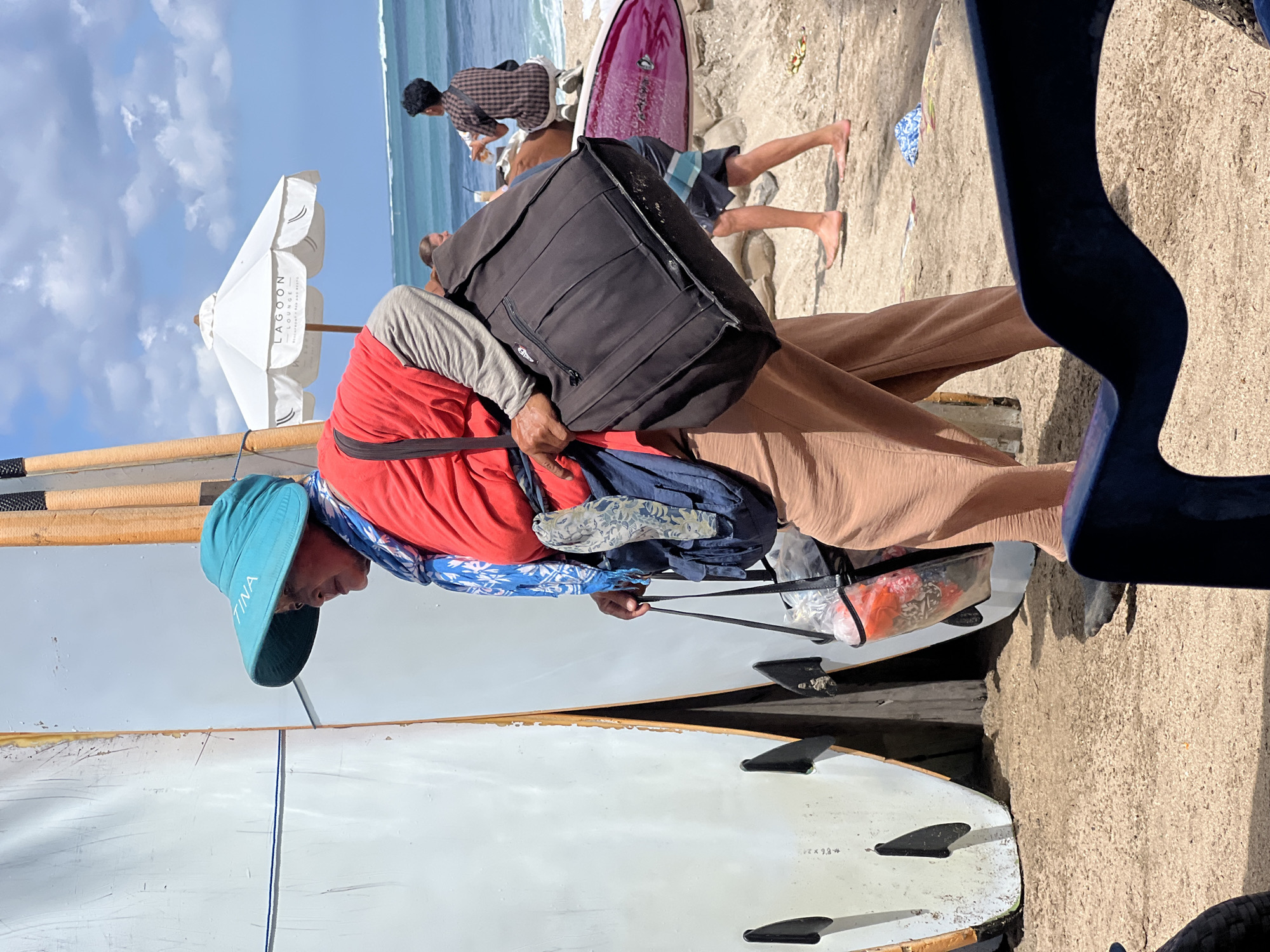 In Kuta, Indonesia, on a sunny day, a person wearing a turquoise sun hat and a red shirt with gray sleeves, walks past two white surfboards. The person is carrying a large black bag over their left shoulder and holding a clear plastic bag containing items with red and floral patterns in their right hand. They are also wearing a blue and white patterned scarf around their neck and loose-fitting, tan pants. The background shows a sandy beach with people in the distance, the ocean with waves, and a white umbrella with the text "LAGOON LOUNGE" printed on it. Other surfboards, including a purple one, are visible on the sand. The scene appears to be a beachside location, possibly a surf rental or beach club area.
