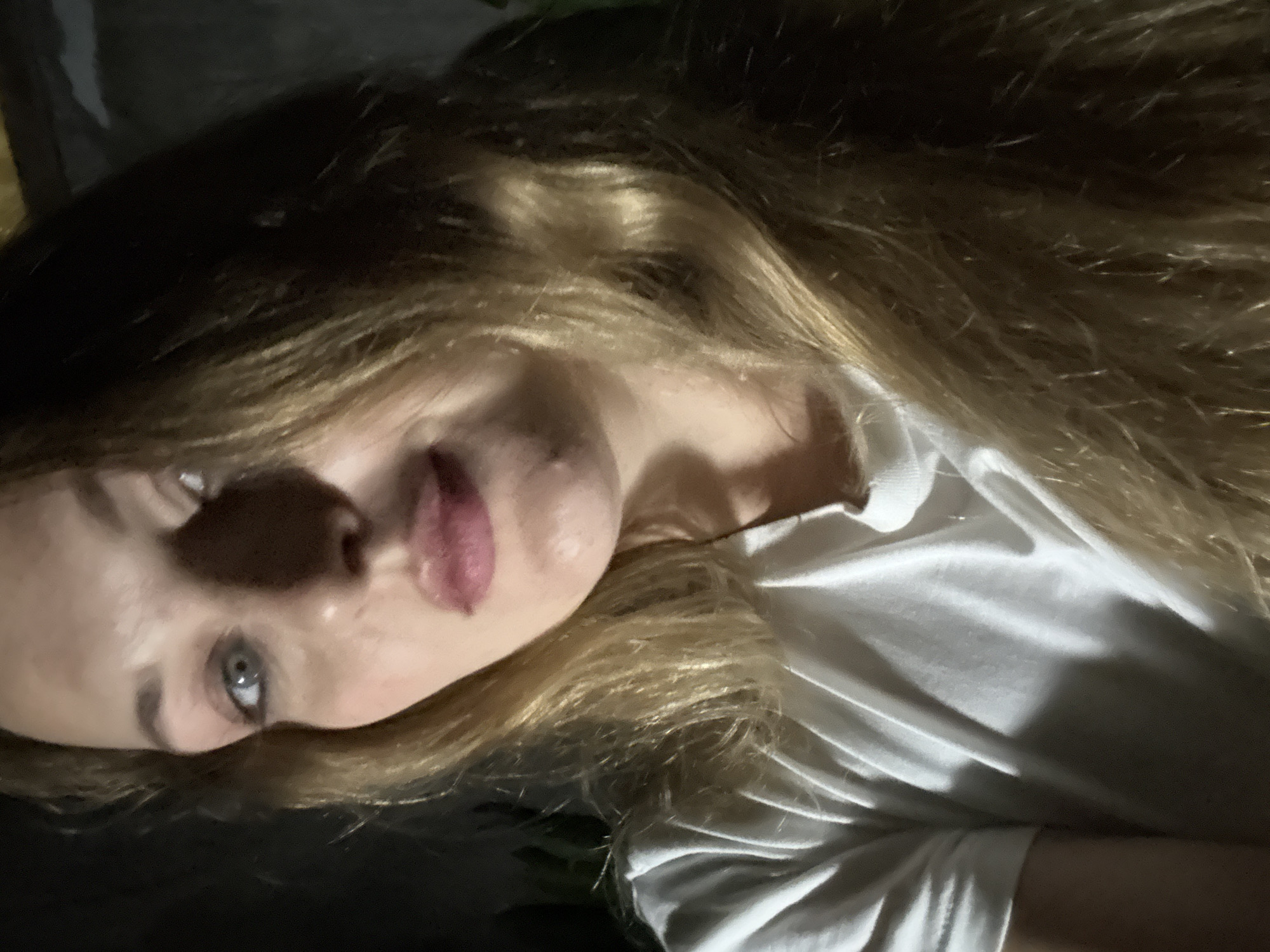 A woman with long blonde hair and blue eyes is shown in a close-up, slightly downward-angled selfie. She is wearing a white t-shirt. Her face is partially illuminated, with the left side of her face in shadow. The background is dark and out of focus, suggesting an indoor setting at night. There is no information in the image to indicate the location in Denpasar, Indonesia.