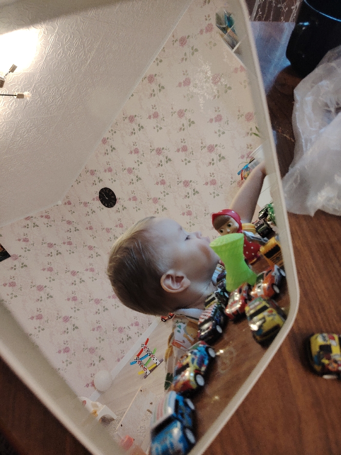 A child's reflection appears in a mirror. The child is looking upwards, surrounded by toy cars and a green toy. The reflection also captures a room with floral wallpaper, a white ceiling with a light fixture, and a black clock. A black object and crumpled white plastic are visible on a wooden surface to the right of the mirror. The mirror has a white frame.
