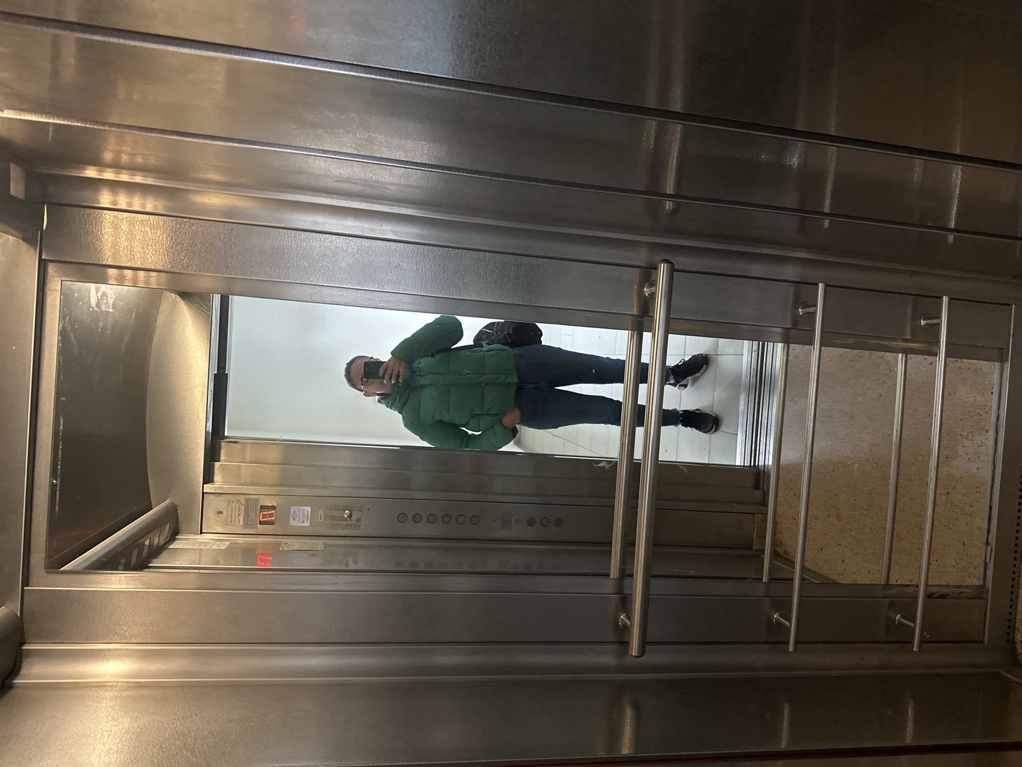A man in a green jacket and black pants is standing inside a metal elevator, taking a picture of himself in the mirror. The elevator has rows of buttons and an illuminated number display showing "11". There is a metal railing in front of the mirror, and the floor appears to be tiled. The location is possibly in Sofia, Bulgaria, but there's no explicit information in the picture to confirm it.
