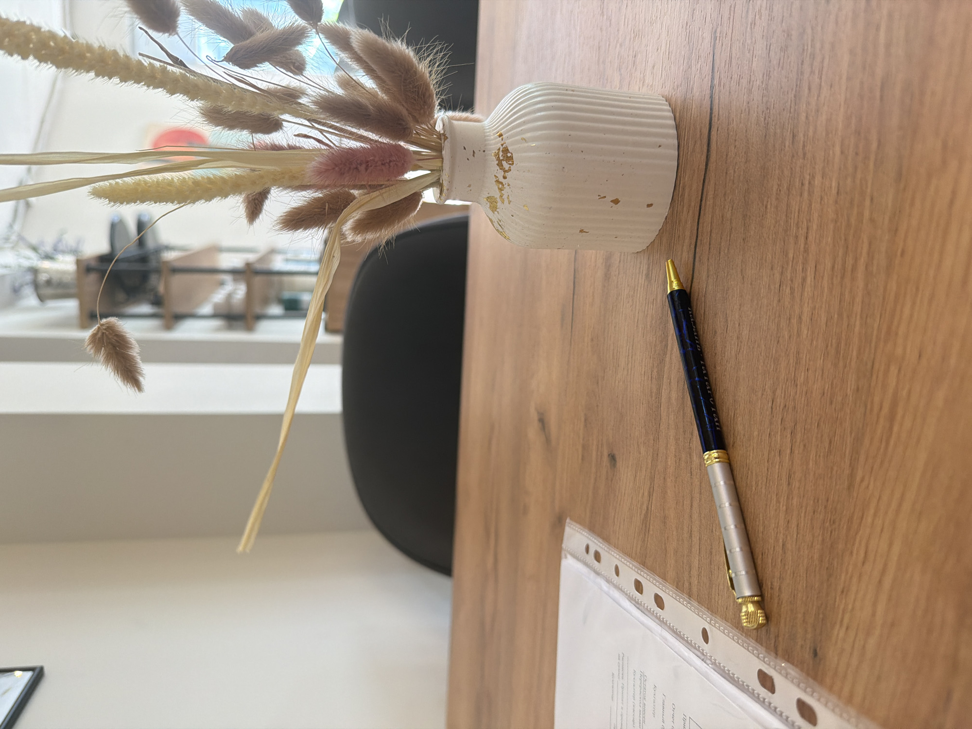 This close-up, rotated image captures a still life arrangement on a light brown wooden desk or table, likely in an office setting in Sochi, Russia.
In the foreground, slightly to the left, is a white, ribbed ceramic vase adorned with gold flecks. It holds a bouquet of dried grasses and flowers in various earthy tones, including light beige, sandy brown, and hints of pale pink. These appear to be types of pampas grass or bunny tail grass.
To the right of the vase, positioned diagonally across the wooden surface, lies a dark blue Pelikan Souverän pen. It features a silver-toned middle section and prominent gold accents on its tip and clip.
Partially visible in the bottom right corner is a document secured within a clear, perforated plastic sheet protector. Visible Cyrillic text on the document includes headings such as "Отчет о проделанной работе" (Report on work performed), "Отдел по работе с клиентами" (Customer Relations Department), "ГЛАВНЫЙ БУХГАЛТЕР" (Chief Accountant), "Бухгалтер" (Accountant), and "Директор" (Director), indicating an administrative or corporate context.
The background is softly blurred, showing a bright interior space with natural light. Elements like a dark-colored chair and a light-colored shelf or counter are vaguely discernible, suggesting an office, reception area, or similar professional environment. The time of day appears to be daytime due to the ambient light. No specific activity is taking place, and no emotions are conveyed.