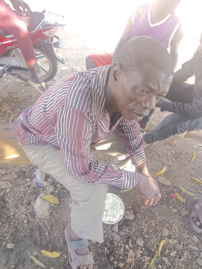 An elder sits on a makeshift bench outdoors. He wears a striped shirt and beige pants with sandals. A white bowl, seemingly filled with a starchy food, rests on the ground before him. There is a motorbike parked nearby and people sitting and standing behind him. The ground is dirt with rocks and a few scattered leaves. The light is bright, casting strong shadows.