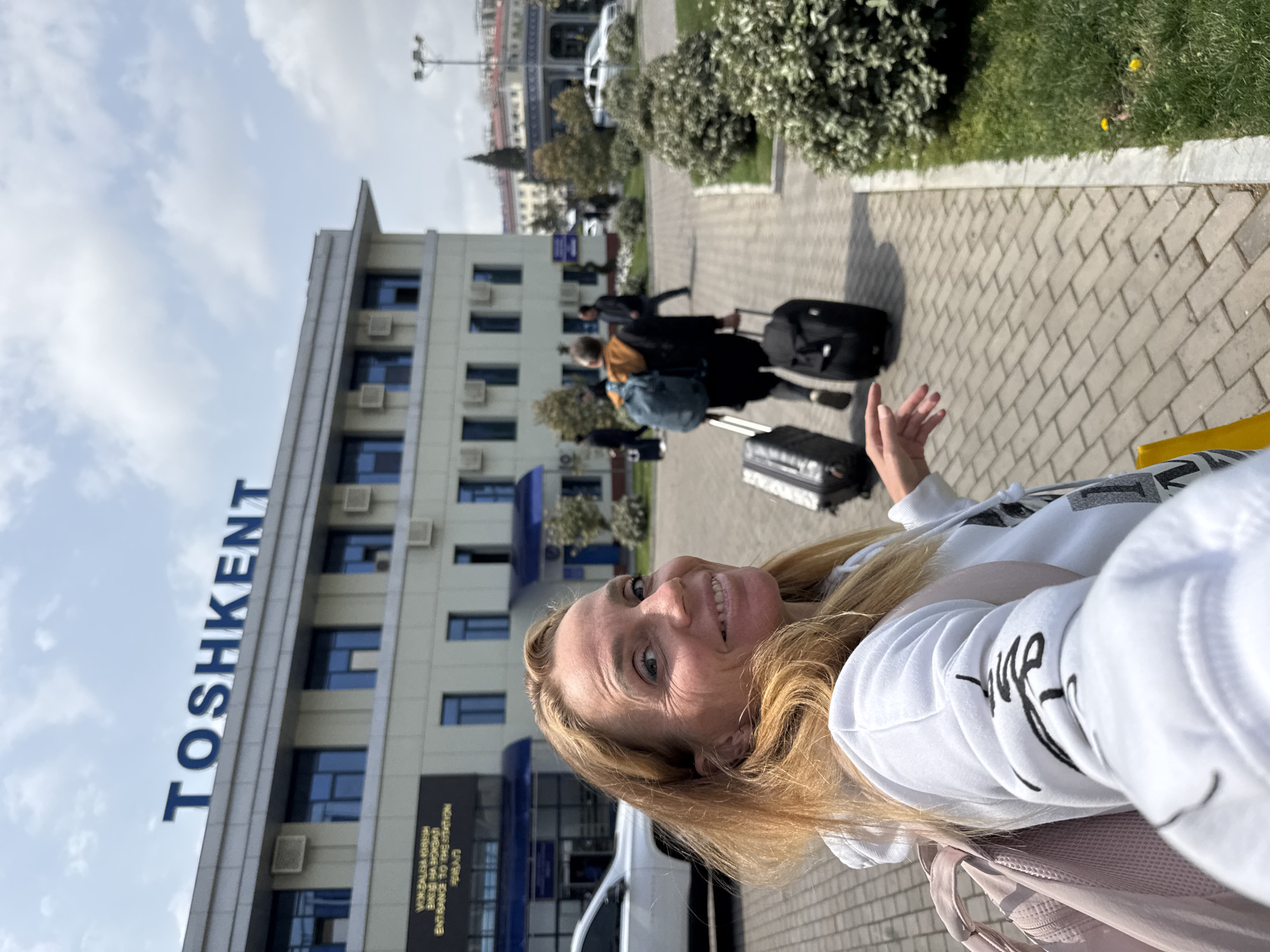 A woman is standing on a brick sidewalk in Tashkent, Uzbekistan, in front of a building with the word "TOSHKENT" on its facade. Behind her, a few people are walking with suitcases. The sidewalk is bordered by green grass and bushes. A white car is parked near the building entrance. The sky is partly cloudy. The woman is wearing a white sweatshirt and a backpack.