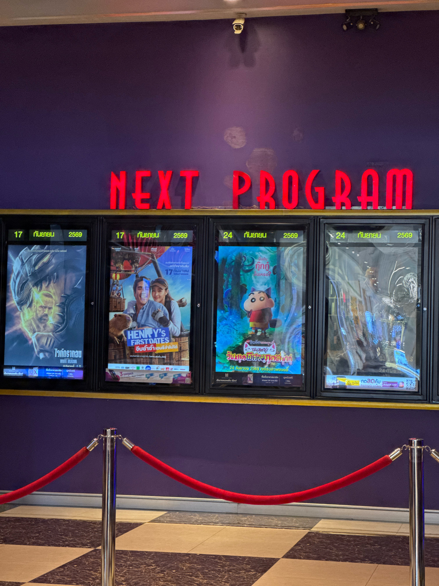 The image shows a movie theater lobby in Samut Prakan, Thailand. The word "NEXT PROGRAM" is displayed in large red neon letters on a purple wall. Below the neon sign, there are four movie posters displayed in illuminated frames. Red velvet ropes on stanchions are set up in front of the display. The floor is tiled in a black and white checkered pattern. The movie posters advertise the following films: a superhero movie, "Henry's First Dates," a Shinchan movie, and another film that is difficult to discern due to reflections. Dates displayed on the posters include September 17 and September 24, 2569 (Buddhist Era), which corresponds to 2026 AD.