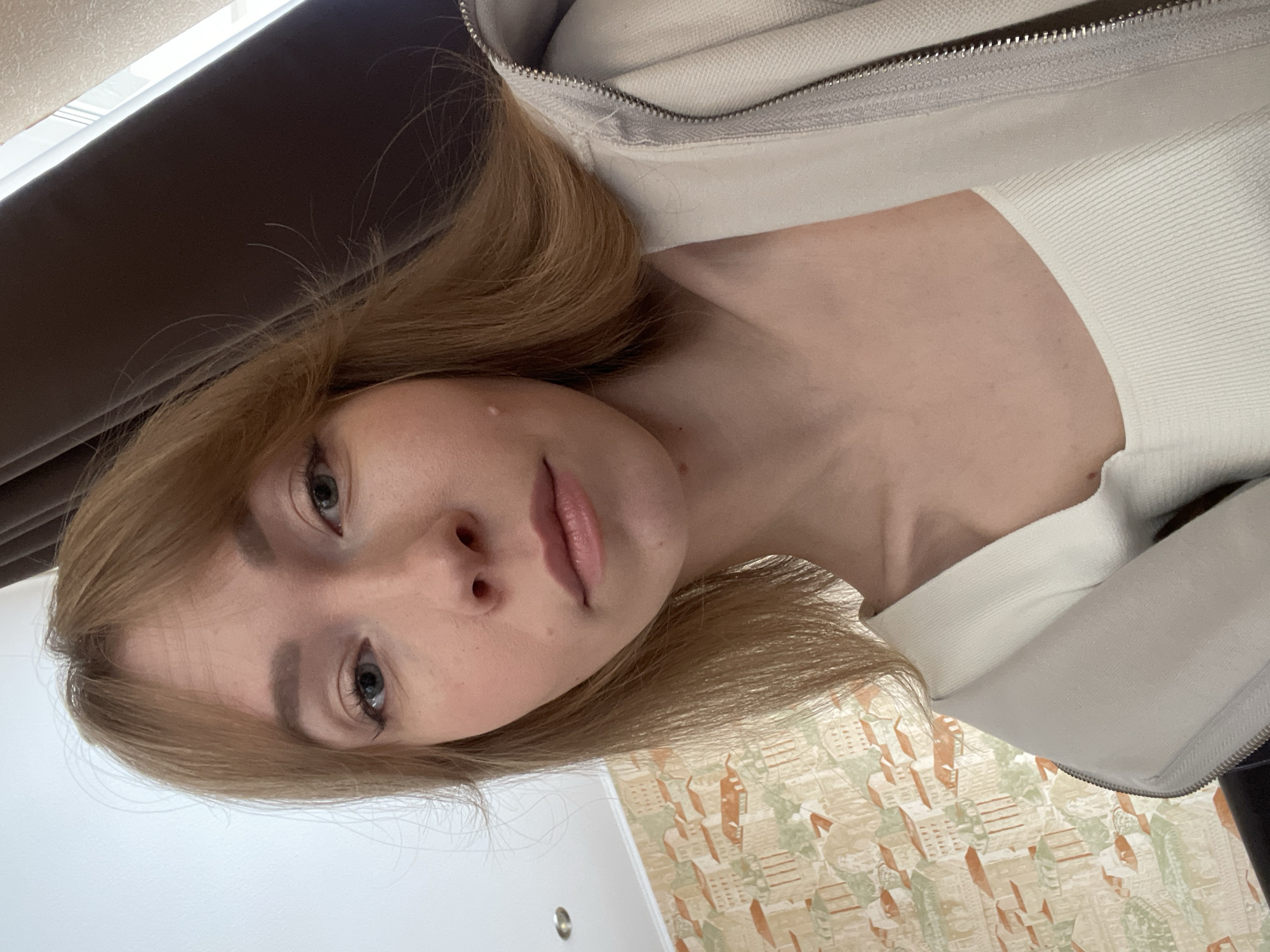 A self-portrait features a young woman with light blonde or light brown hair, captured from a tilted, slightly upward-looking angle. She is looking directly towards the camera with a neutral and calm expression. Her eyes are a light blue or grey color, and her lips are a natural light pink. She appears to be wearing a light-colored, possibly off-white, ribbed top, layered under a beige or light grey zip-up jacket with a visible zipper detail around the collar and shoulder.
The background reveals interior elements. To the upper left of the frame, a dark-colored curtain or window blind is partially visible, suggesting a window and natural light source. Below her head and extending to the lower right, a wall is adorned with patterned wallpaper. This wallpaper depicts a repeating design of stylized buildings or houses in shades of orange, green, and beige on a lighter background. A plain, light-colored wall or ceiling is visible in the bottom-left corner, providing a simple contrast. The overall lighting appears to be soft and natural, indicating daytime. The scene takes place indoors, likely in a room within a residence in Perm, Russia. There is no visible text in the image.