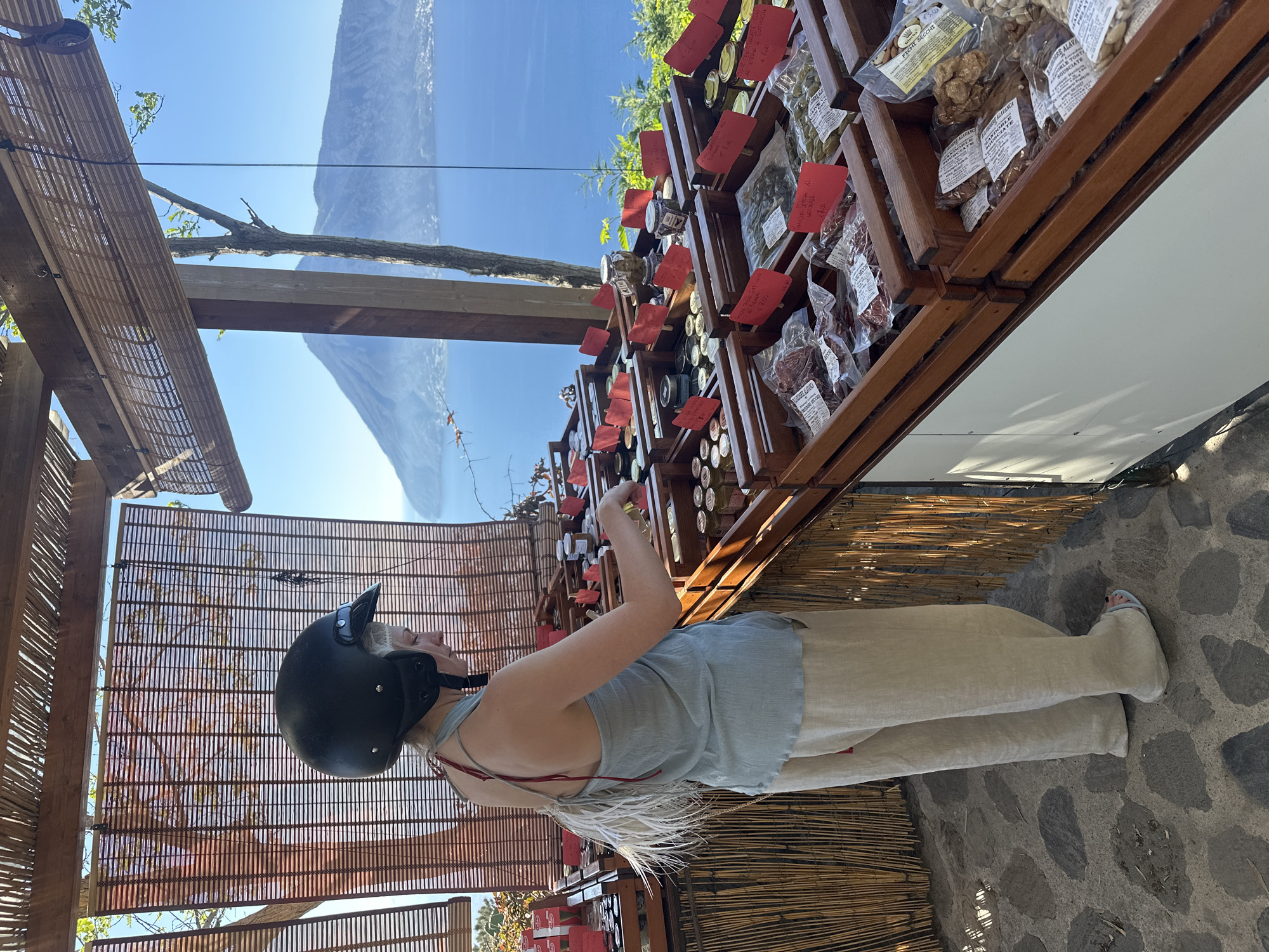 The image, rotated 90 degrees clockwise, presents an outdoor market scene under a bright, clear blue sky in what appears to be Lipari, Italy. A woman, seen from her side and back, stands at a rustic wooden market stall, engaged in browsing or selecting products. She wears a light grey sleeveless top, light-colored long pants, and light blue sandals, with a black motorcycle helmet on her head. White fringed elements are visible near her back, possibly part of her attire or a bag.
The market stall is made of dark wood, featuring shelves laden with various packaged goods, including items in clear plastic bags and glass jars. Numerous red labels with handwritten prices and product names are affixed to the shelves and items. Visible Italian text includes "Fichi Secchi" (dried figs), "Mandorle" (almonds), "Nocciole" (hazelnuts), "Pistacchi" (pistachios), "Miele" (honey), "Pomodori Secchi" (dried tomatoes), and "Origano" (oregano), with prices such as "€ 2.50" and "€ 4.00."
In the background, a breathtaking mountainous landscape stretches under the bright blue sky, indicative of an island or coastal setting overlooking the sea. The foreground shows a textured stone pavement and a woven bamboo canopy structure, suggesting an open-air venue designed for shade. The scene depicts a sunny daytime activity, highlighting local produce against a scenic natural backdrop.