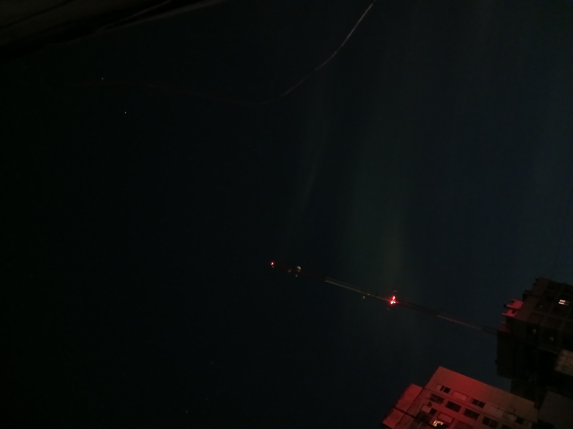 The image shows a nighttime scene with a dark sky and several buildings illuminated by red and white lights. In the lower right corner are two buildings, one appearing with red-tinted windows and the other darker, with some white-lit windows. A crane is visible, stretching across the sky between the buildings. The sky itself is very dark, with some faint vertical streaks. A thin cable is seen in the upper part of the picture. Due to the prominent presence of buildings and a construction crane, it is likely that the location is in the city of Buenos Aires, Argentina.